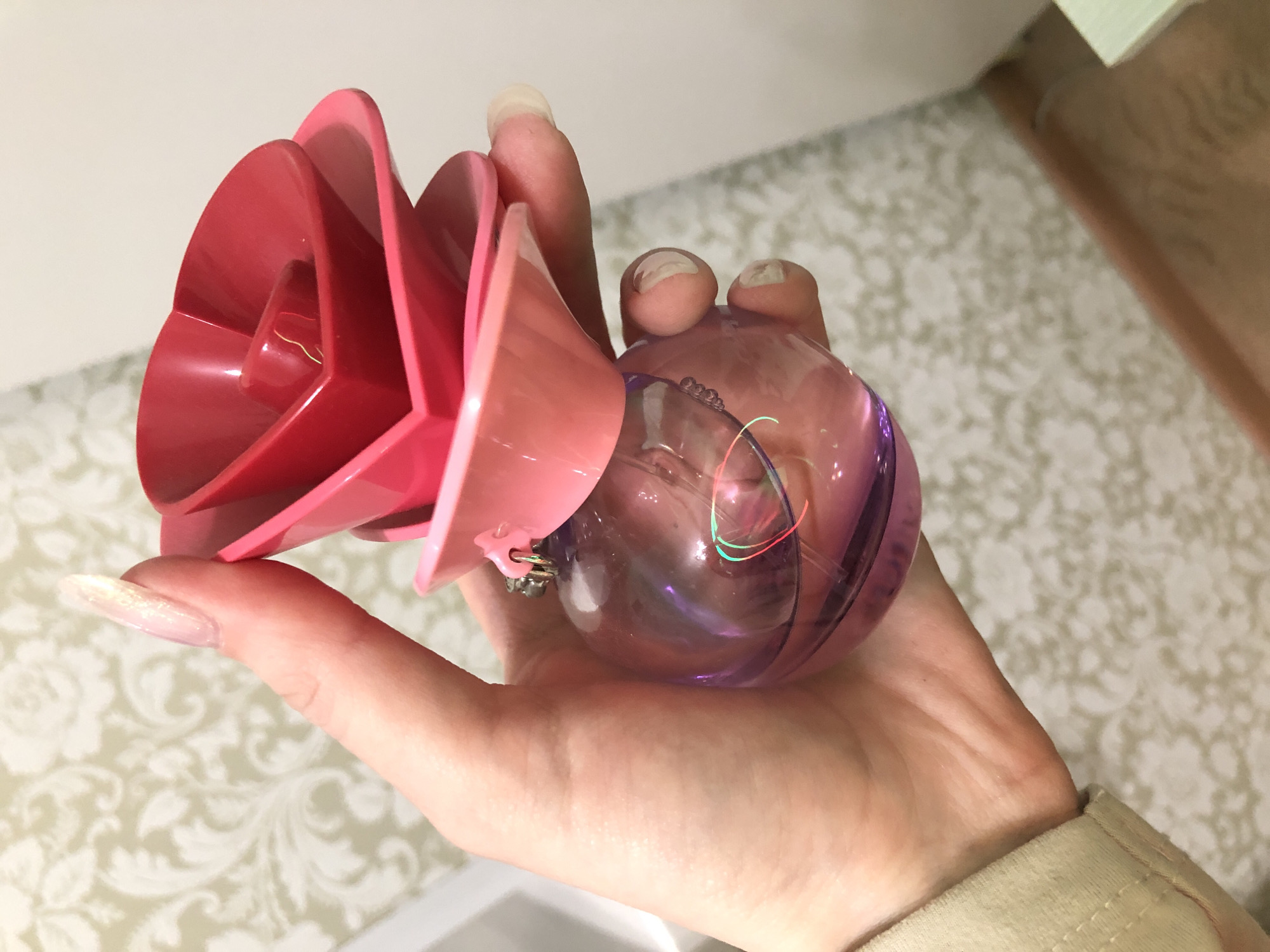 A right hand holds a rose-shaped object. The "rose" has concentric, red-colored plastic layers forming its petals. The base of the rose connects to a spherical, translucent object with an inner coiled wire. The hand has long nails. The background is a wall with a patterned wallpaper.