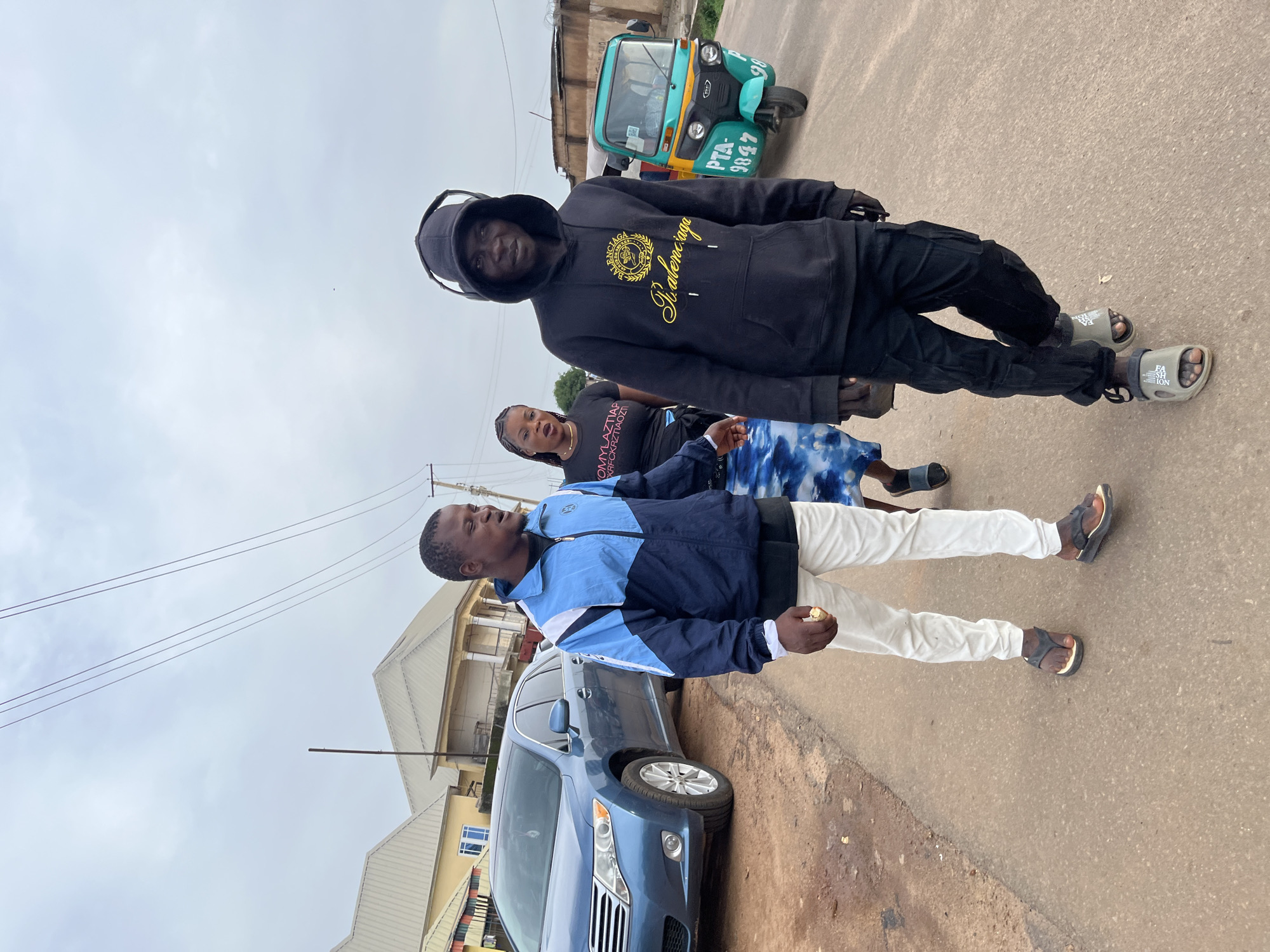 The image, taken in Gura Top, Nigeria, shows a group of people on a street with buildings and a vehicle visible. In the foreground, a young man wearing a blue and white jacket, white pants, and dark sandals is walking and looking to his left. He is holding a piece of food in his right hand. Behind him, to his right, a young woman in a black t-shirt with white text and a blue and white tie-dye skirt is also walking. Further back and to the left, another young man is standing, wearing a black hoodie with gold lettering and dark pants and sandals. He is looking towards the camera. To the far right of the frame, a green and yellow auto-rickshaw is parked on the street. The sky is overcast, suggesting it could be daytime, possibly late morning or early afternoon. The street appears to be paved, with some patches of dirt and debris. Buildings with light-colored roofs are visible in the background. The overall atmosphere appears casual, with the individuals seemingly engaged in everyday activities. Some text is visible on the auto-rickshaw: "PTA-9847". The lettering on the woman's t-shirt is partially obscured but appears to be in white. The hoodie worn by the man in the foreground has gold text that reads "Balenciaga".