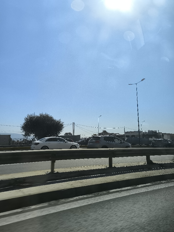 The image, taken from a car window, shows a road with several vehicles moving in the opposite direction. The car is driving under a highway overpass, with a metal guardrail visible in the foreground. The sun is shining brightly in a clear blue sky, though there are some light, blurry clouds. Trees are visible in the distance, as well as a building with the word "SMIT" partially visible on a sign. There is a large tree in the middle of the road, and a few streetlights are visible against the sky. The image is taken in the city of Egaleo, Greece, at coordinates 37.98 latitude, 23.68 longitude.