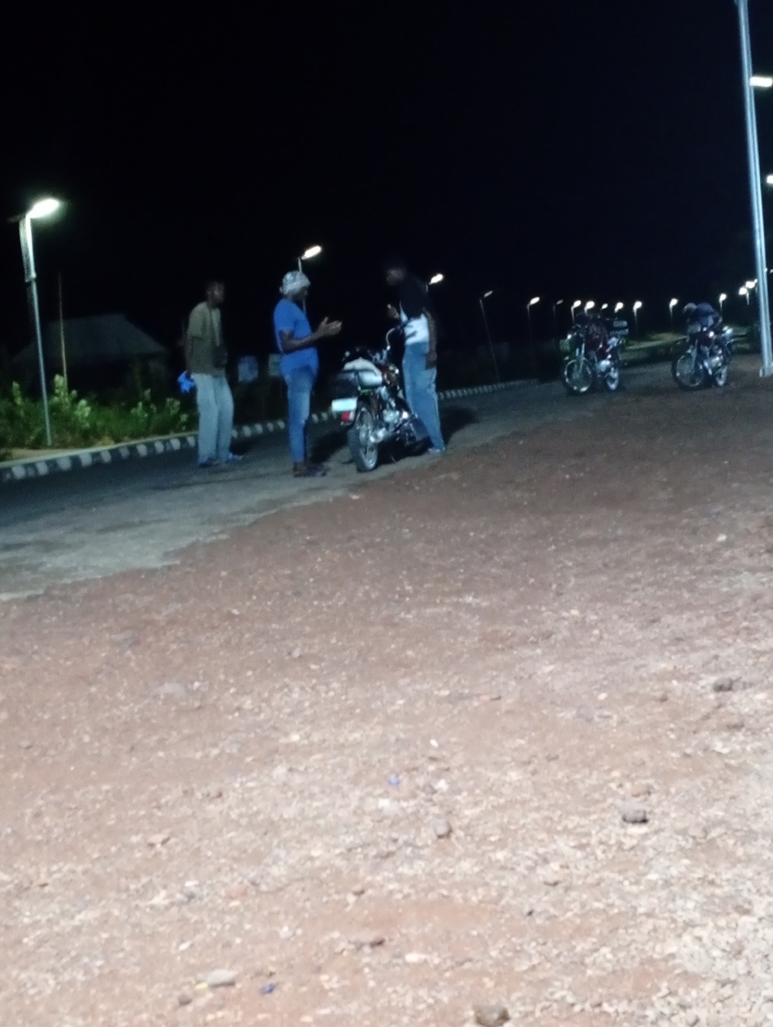 The image depicts a night scene in Adankolo, Nigeria. Three individuals are present on a paved surface adjacent to a dirt area. One person wears a light-colored shirt and darker pants, another wears a blue shirt and blue pants, and the third is wearing a dark shirt and light pants. They are gathered around a motorcycle. Two other motorcycles are parked further down the paved area. Streetlights illuminate the scene. A low building and some vegetation are visible in the background. The sky is dark.