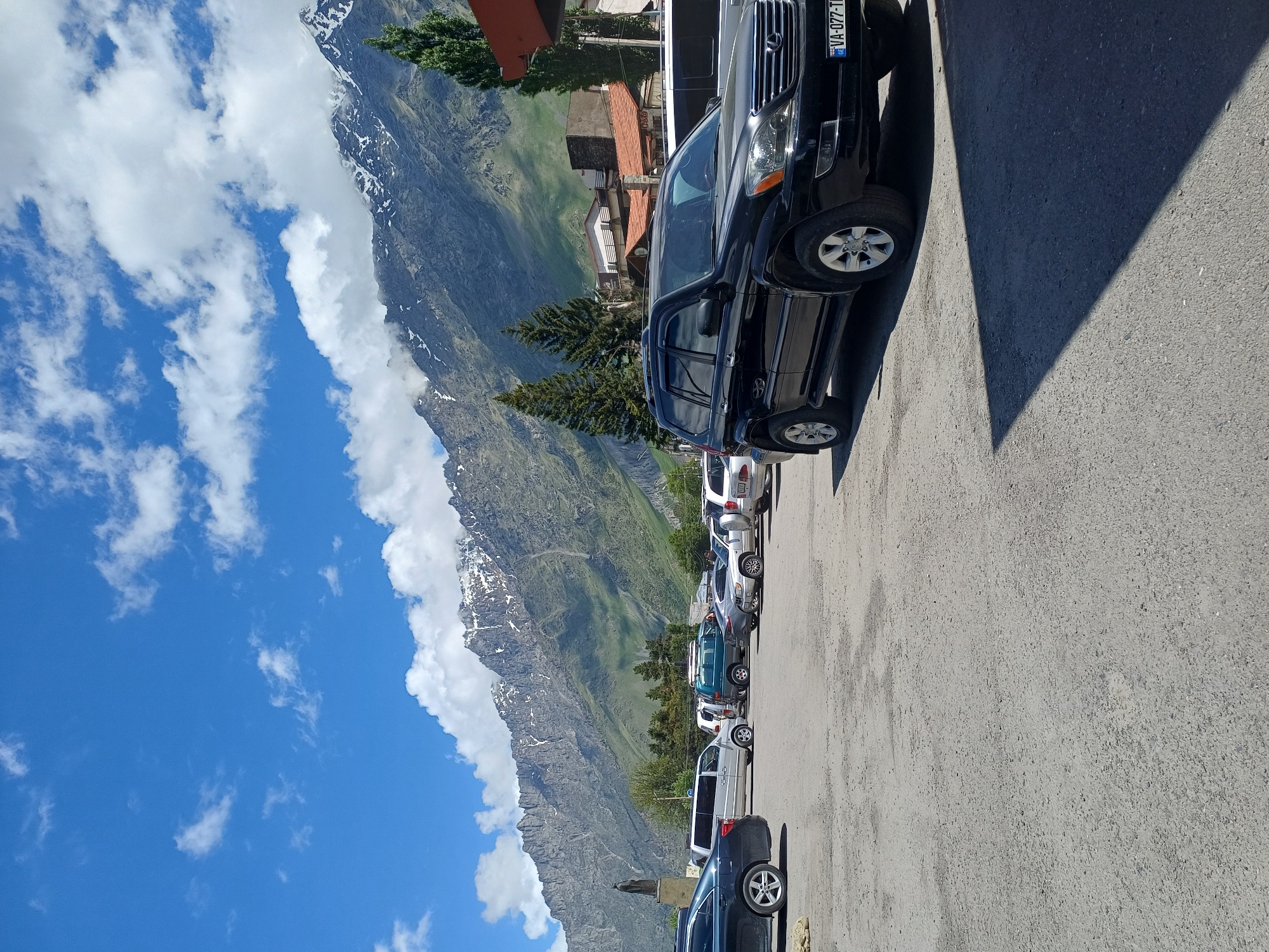 A black Lexus SUV is parked in a lot in front of a backdrop of a majestic mountain range. The SUV is positioned in the foreground, facing the viewer's left. Behind it, other cars and vans are parked. The mountains in the distance are rugged and covered with snow at their peaks. The sky is a vibrant blue with scattered white clouds. The scene appears to be in a town, possibly a tourist destination, given the presence of multiple vehicles and the scenic mountain views. The license plate of the Lexus is VA-077-TI. The image is taken during the daytime with good lighting.