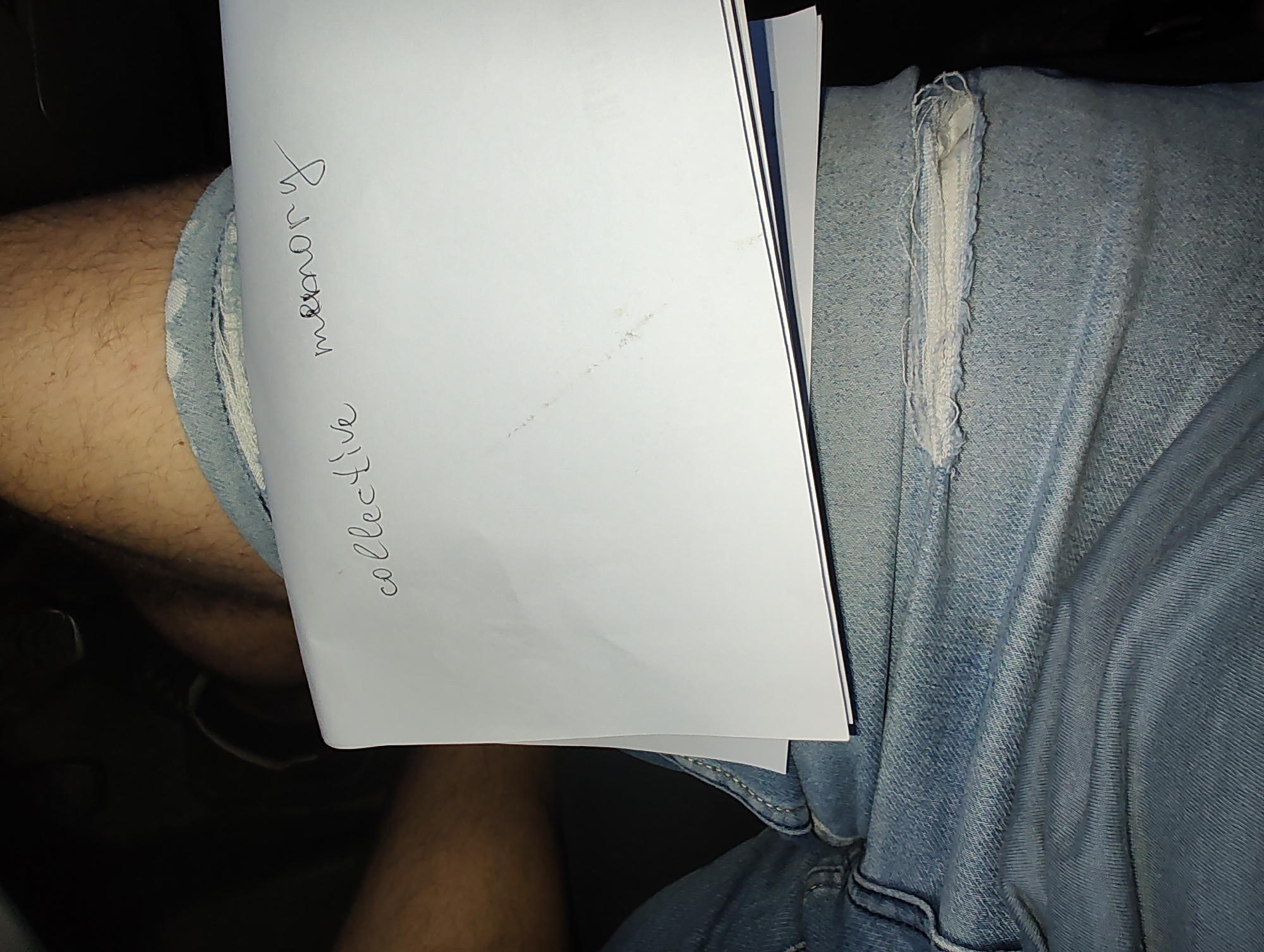 The image shows a close-up view of a person's lap area, featuring denim jeans with a distressed section. A white sheet of paper is held across the lap, with handwritten text in black ink visible on its surface. The text reads "collective memory." The environment appears to be indoors, with dark shadows suggesting a dimly lit setting. No specific location in Eutin, Germany, can be identified from the visual information provided in the image.