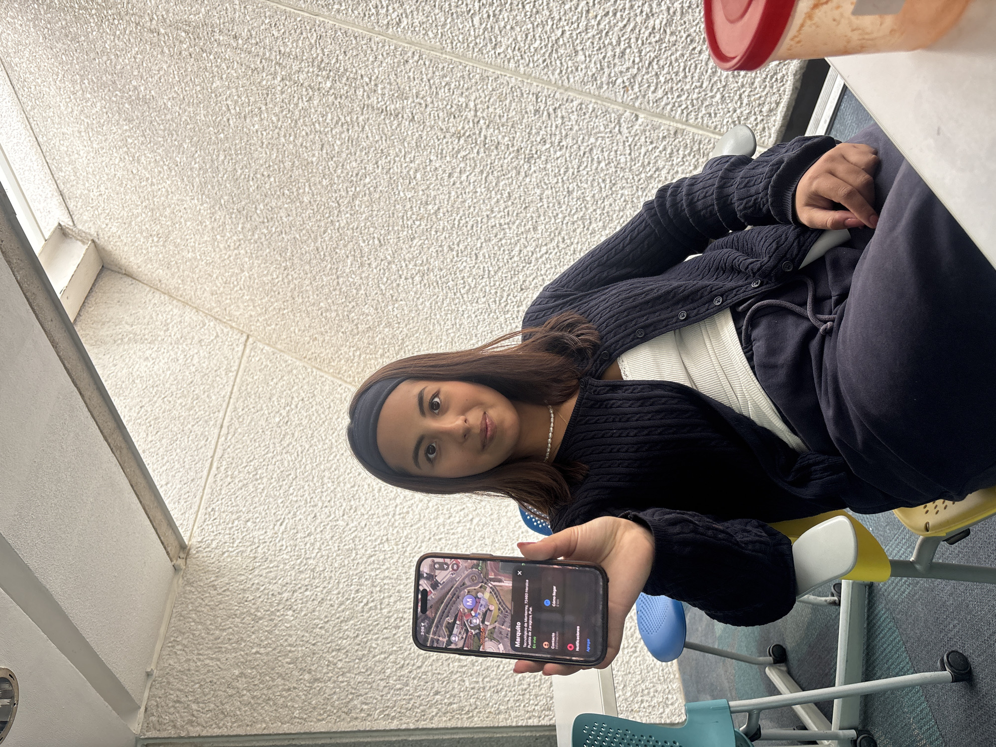 A young woman is prominently featured, seated and holding up a smartphone toward the viewer. She has long dark hair, a dark headband, and wears a black knitted cardigan over a white top, paired with dark pants. Her expression is neutral as she looks directly into the camera.
Her right hand extends the smartphone, displaying a map application interface. The screen shows a detailed satellite view with multiple markers and an information panel. Clearly legible text on the phone identifies a location as "Marquito" within "Tecnológico de Monterrey, 72460 Heroica Puebla de Zaragoza, Pue." Additional options like "En vivo," "Contacto," "Cómo llegar," "Notificaciones," and "Agregar" are also visible. The phone's status bar indicates the time as 3:00.
The setting appears to be an indoor or covered outdoor area, possibly a university common space or cafeteria. The background features a textured, light-colored wall. A white table is visible to the woman's right, topped with a container that has a red lid. The floor is covered with multi-colored carpet tiles in blue, yellow, and teal patterns. The chairs, visible around her, are made of plastic in matching bright colors, featuring metal frames and wheels, typical of educational or casual seating. The scene suggests a moment captured at the Tecnológico de Monterrey campus in Heroica Puebla de Zaragoza, Mexico.