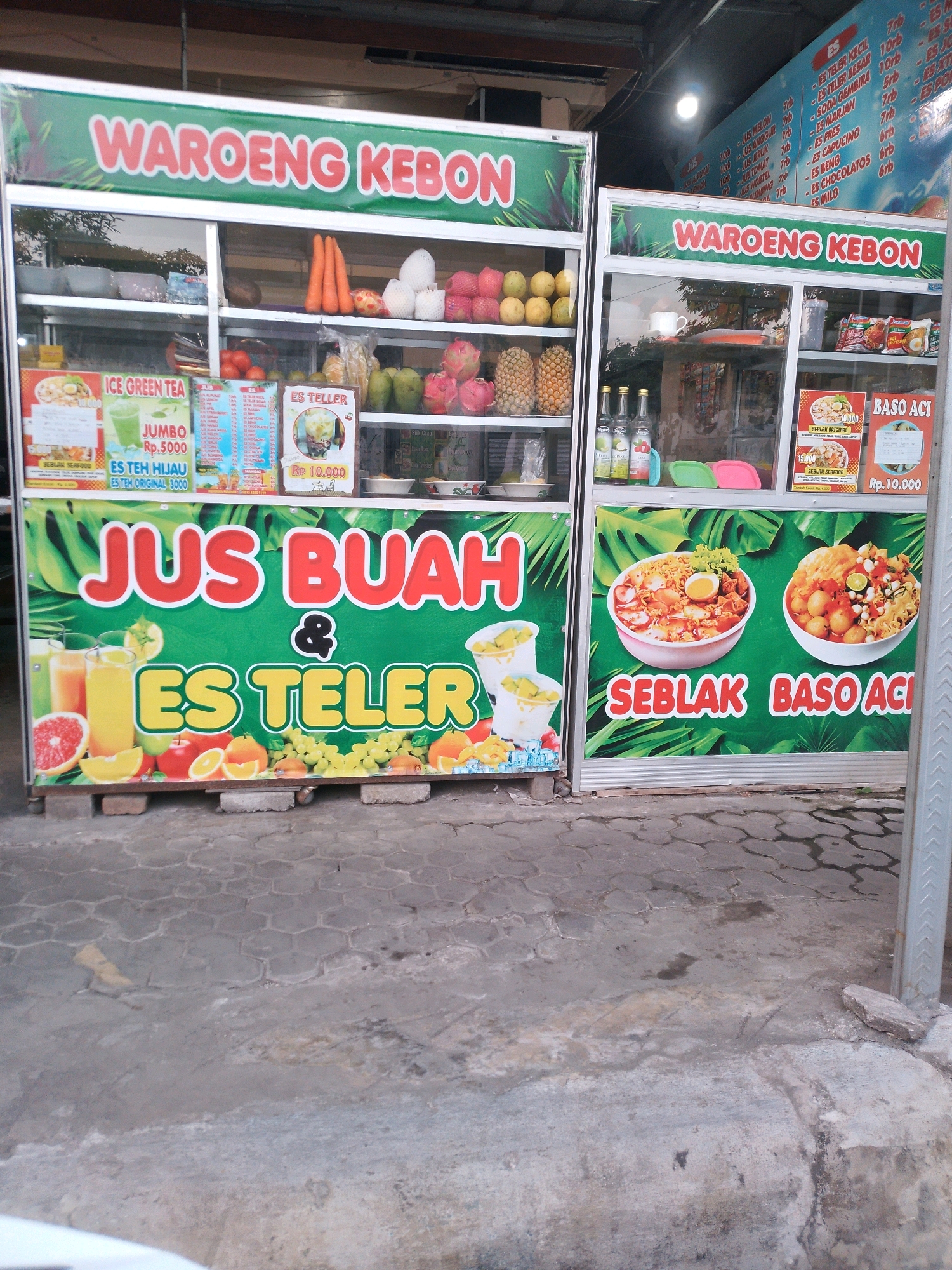 This is a medium shot, slightly angled photograph of two food stalls in Kertosono, Indonesia. The stalls, branded "WAROENG KEBON," sell fruit juices, iced desserts, and seblak (a spicy Indonesian dish). The stalls are constructed of metal and glass, with colorful signage and displays of food and drinks.
The stall on the left features a sign that reads "JUS BUAH & ES TEL ER" (Fruit Juice & Iced Tel ER). Below this are images of glasses of juice and bowls of iced dessert. The display also includes various fruits such as grapefruit, oranges, apples, and grapes. Behind the glass, there are bottles of what appear to be syrup or juice, and some packaged food items.
The stall on the right has signage for "SEBLAK BASO ACI" (Seblak Meatball Aci). Two bowls of seblak are depicted on the sign. Behind the glass, there are more packaged food items and some drinks.
The stalls are situated on a paved area with a hexagonal pattern, typical of sidewalks or public spaces. The background shows a building with a corrugated metal roof, suggesting a semi-open-air environment. The overall impression is that of a bustling street-side food vendor. There are no people visible in the photograph.
