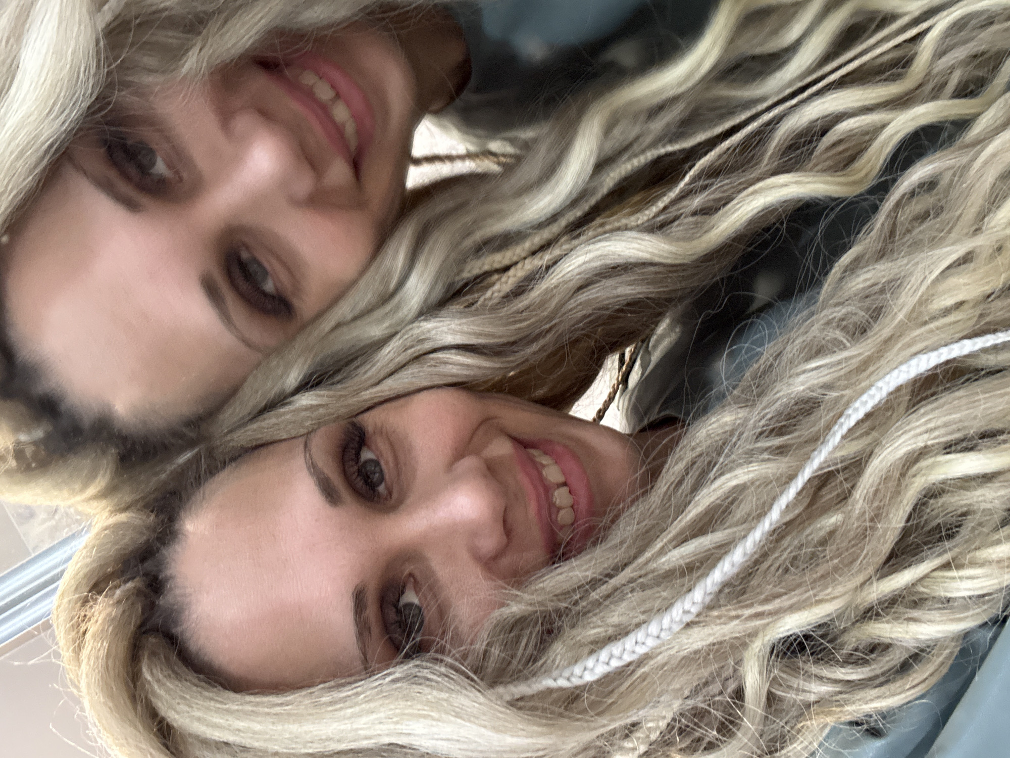 A close-up, selfie-style image features a woman with long, blonde, wavy hair. The woman is smiling, revealing her teeth. The image is presented at an angle, suggesting it may have been taken in a mirror. The location is not discernible from the image.