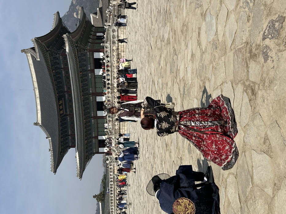 The image depicts an outdoor scene within the Gyeongbokgung Palace complex in Seoul, South Korea, with the prominent Geunjeongjeon Hall in the background. The day is clear and sunny with a blue sky.
In the foreground, a person with short, reddish-brown hair, wearing a black top with gold embroidery and a flowing red and black patterned skirt (resembling a modern hanbok), is crouched low, holding a smartphone or camera to take a photograph. To their left, another individual is kneeling, facing away from the camera, dressed in a dark blue traditional Korean robe with a circular gold emblem on the back and a black traditional hat, paired with modern black sneakers.
In the mid-ground, a person with reddish-brown hair and glasses stands centrally, facing the camera, posing for the photograph. This individual wears a white inner garment beneath a patterned, vest-like outer layer and a small black traditional headpiece.
The background is dominated by the large, multi-tiered Geunjeongjeon Hall, characterized by its dark tiled roofs and intricate traditional Korean architecture. A considerable crowd of people is visible on the steps and raised platform of the hall. The extensive courtyard is paved with irregular stone slabs. Scattered across the courtyard are numerous other individuals, many of whom are dressed in various traditional Korean hanbok, engaged in walking, sightseeing, and posing for photographs. Distant mountains are visible on the horizon behind the palace structures.