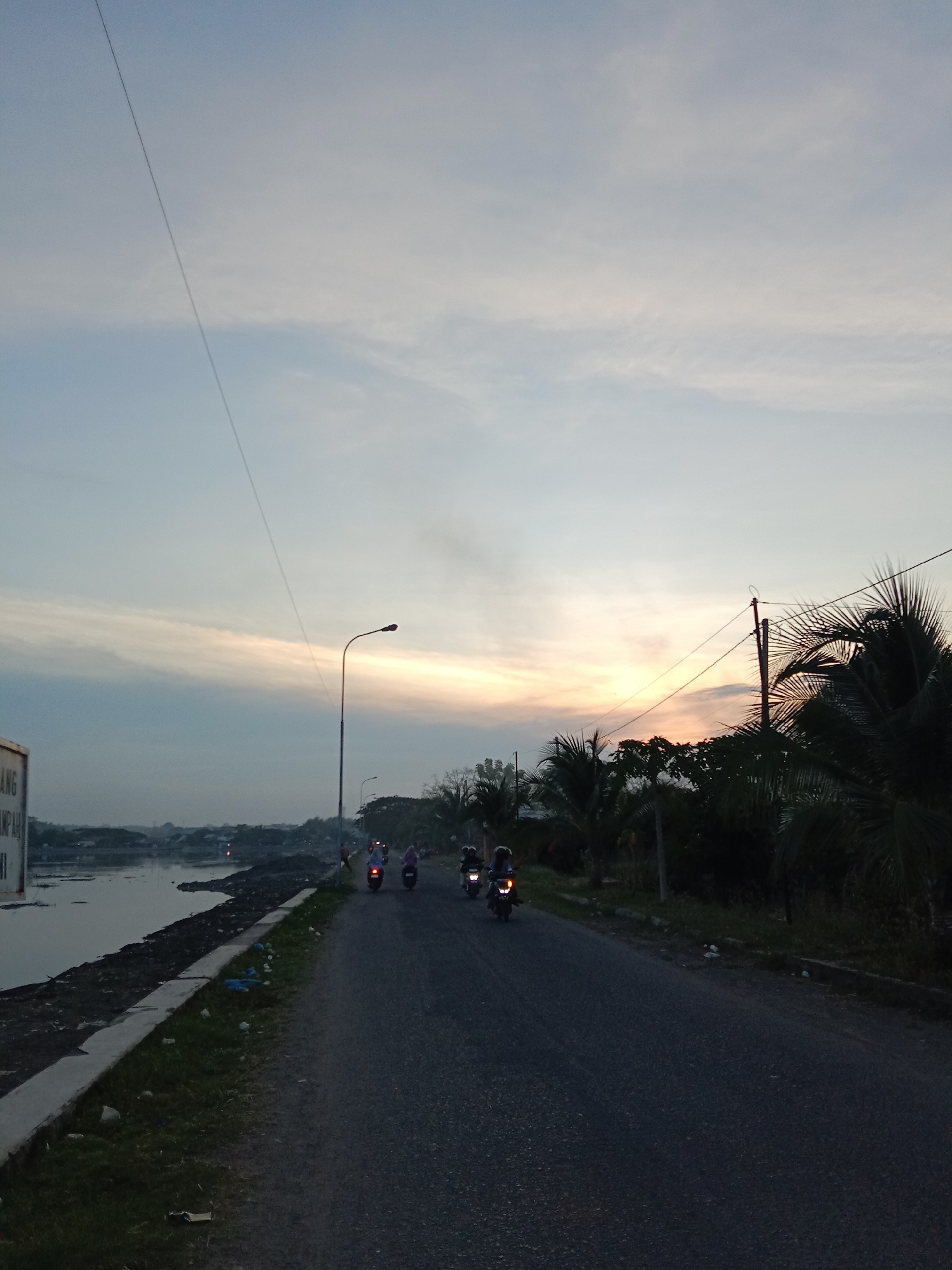 The image captures an outdoor scene at either dawn or dusk in Lhokseumawe, Indonesia. The sky displays a soft gradient of colors, transitioning from pale blue overhead to warm oranges and yellows near the horizon, indicating the sun's low position. Wispy clouds are scattered across the sky, and a thin wire stretches diagonally from the top-left corner downwards.
In the midground, an asphalt road runs from the lower-center towards the distant horizon. Several people are riding motorbikes on this road; some motorbikes have their headlights illuminated, suggesting low light conditions. To the right of the road, a dense line of tropical vegetation, primarily palm trees, forms a dark silhouette against the glowing sky. A utility pole with wires is also visible among the trees on the right.
To the left of the road, a calm body of water, possibly a river or large pond, parallels the road. Its bank appears somewhat eroded or covered in dirt, with some visible debris. A concrete curb separates the road from a grassy strip, where some litter, including plastic bags, is noticeable. A street light pole stands by the road on the left. Further left, a white sign is partially visible, displaying the Indonesian words "ANG" and "SAMPAH", the latter meaning "trash" or "garbage". The scene depicts a tranquil moment of daily life, with commuters traveling against the backdrop of a colorful sky.