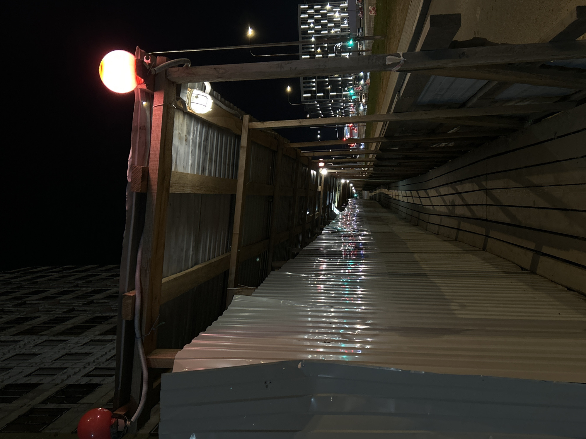 This nighttime image, rotated 90 degrees counter-clockwise, captures a covered pedestrian walkway or temporary tunnel in an urban setting, likely Saint Petersburg, Russia. The dark sky indicates late evening or night, with the scene illuminated by numerous artificial light sources.
In the foreground, a structure of wooden beams and corrugated metal sheeting forms the enclosure. A prominent, bright orange-red spherical light is affixed to a wooden post on the far left. Adjacent to it, a white, cage-like lamp is visible. A white corrugated conduit runs vertically down a wooden support. At the very bottom left, a red hemispherical object is partially visible.
The roof of the walkway, made of reflective corrugated metal, appears wet, glistening with colorful reflections—red, green, blue, and white—that stretch into the distance, suggesting internal lighting or external city lights bouncing off the surface.
Beyond the temporary structure, a paved urban area, possibly a sidewalk or road, is visible. In the background, a tall, modern building with numerous brightly lit, rectangular windows stands against the dark sky. Streetlights with a warm glow and traffic lights showing green and red can also be seen in the distance, indicating an active city environment. No people or visible text are present in the image.