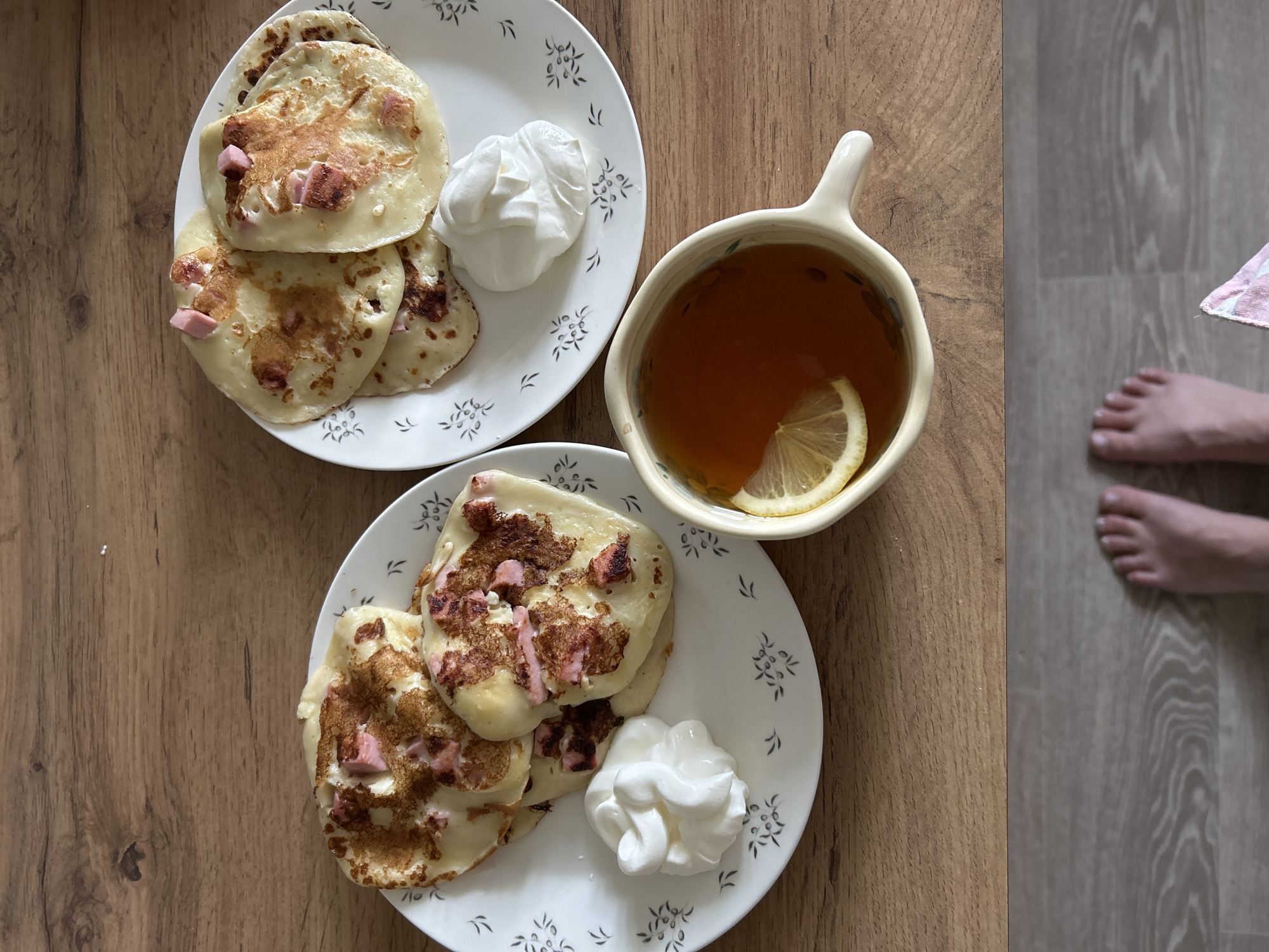 The image shows a still life composition on a wooden surface. Two white plates, each containing three pancakes dotted with small pieces of ham, sit alongside a dollop of whipped cream. A mug of tea with a lemon slice sits next to the plates. A pair of bare feet are visible in the right-hand corner of the image, suggesting a person is present. The scene is set on what appears to be a wooden table or countertop. The location is Chaikovsky, Russia.