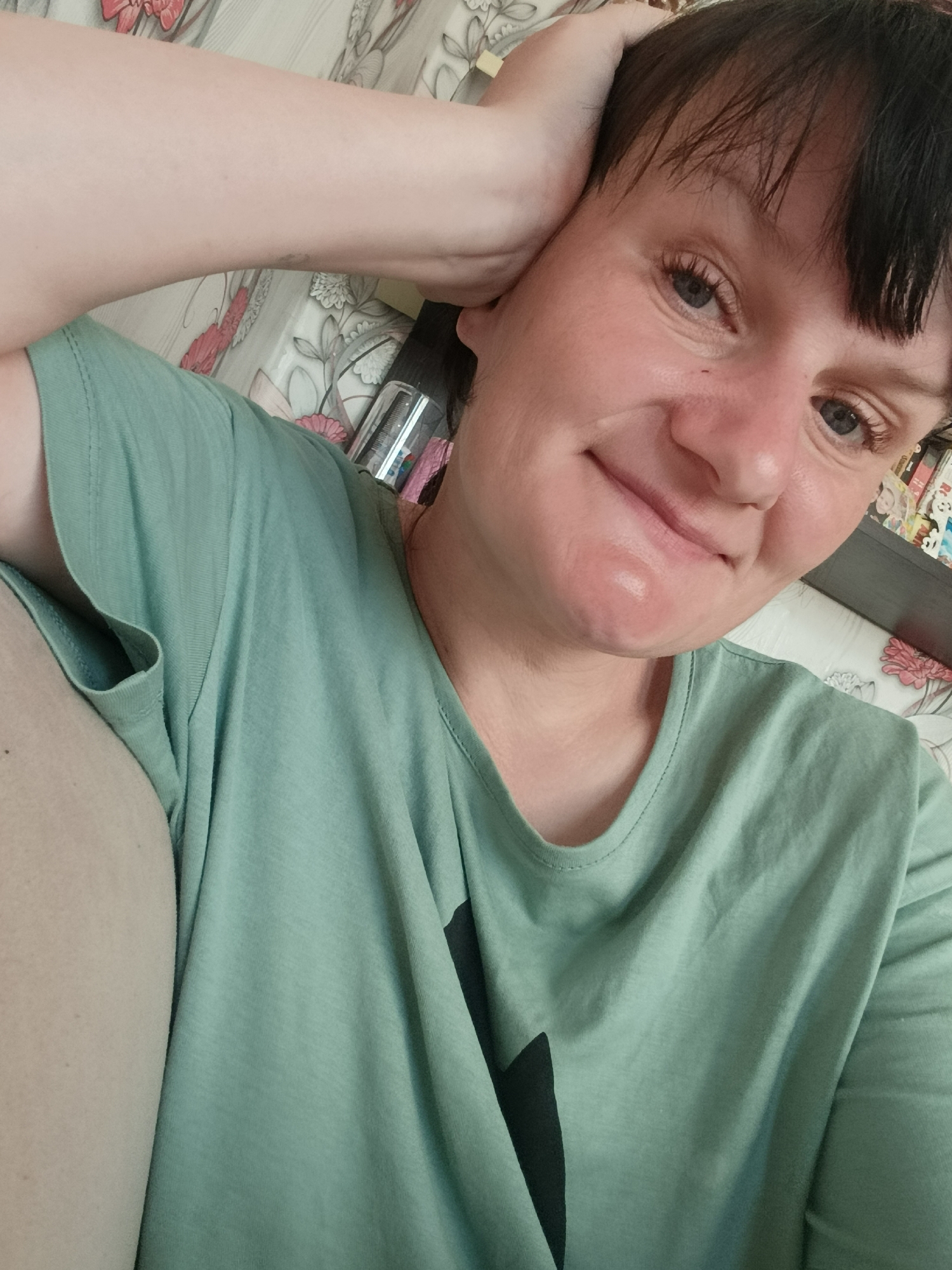 A fair-skinned woman with short dark hair and bangs is posing for a selfie, holding her right hand near her ear. She is wearing a light green t-shirt with a black design on the front. The background appears to be an indoor space with floral patterned wallpaper. Behind her head, there's a shelf with various items.
The location Фоки, Russia cannot be confirmed from the image.