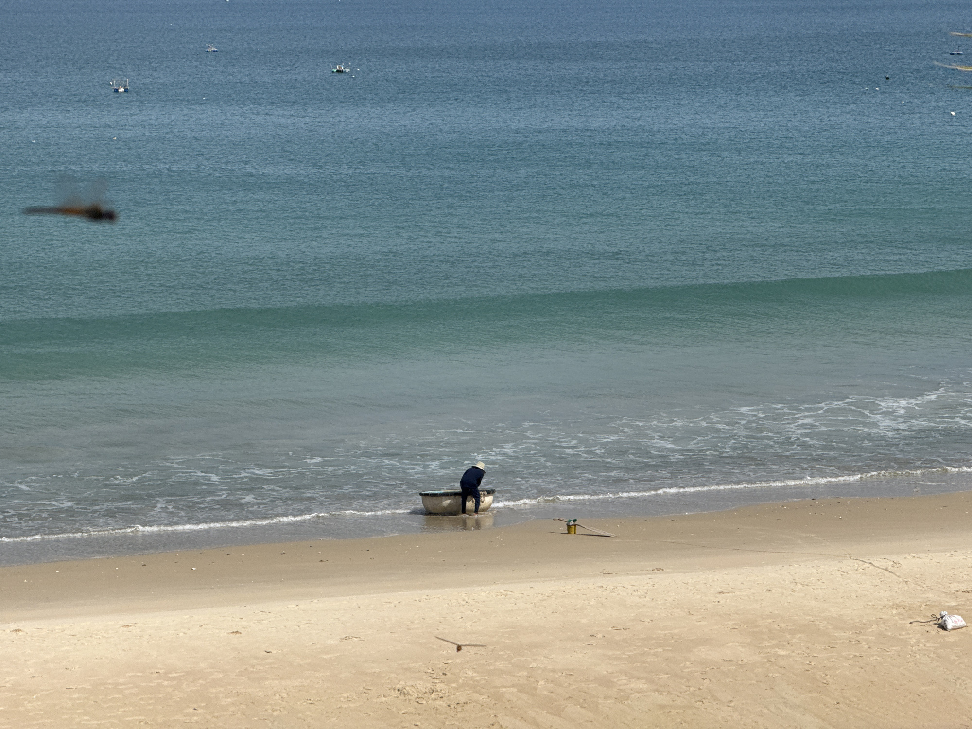 The image depicts a coastal scene with a person interacting with a boat on a sandy beach. The ocean occupies the upper half of the frame, with distinct bands of color indicating varying depths. The water transitions from a deep blue in the distance to a lighter turquoise near the shore, with whitecaps and foam marking the breaking waves. Several small boats are visible further out at sea.
The foreground is dominated by a sandy beach. In the middle ground, a person wearing a dark blue garment and a white hat is bent over, engaged with a small, round boat partially submerged in the shallow water. To the right of the boat, on the sand, are two objects: a yellow bucket and a piece of wood. Further down the beach, towards the right edge of the frame, there is a small, white, bagged object. A shadow of a flying insect is captured in the mid-left of the image. The lighting suggests a bright, sunny day.