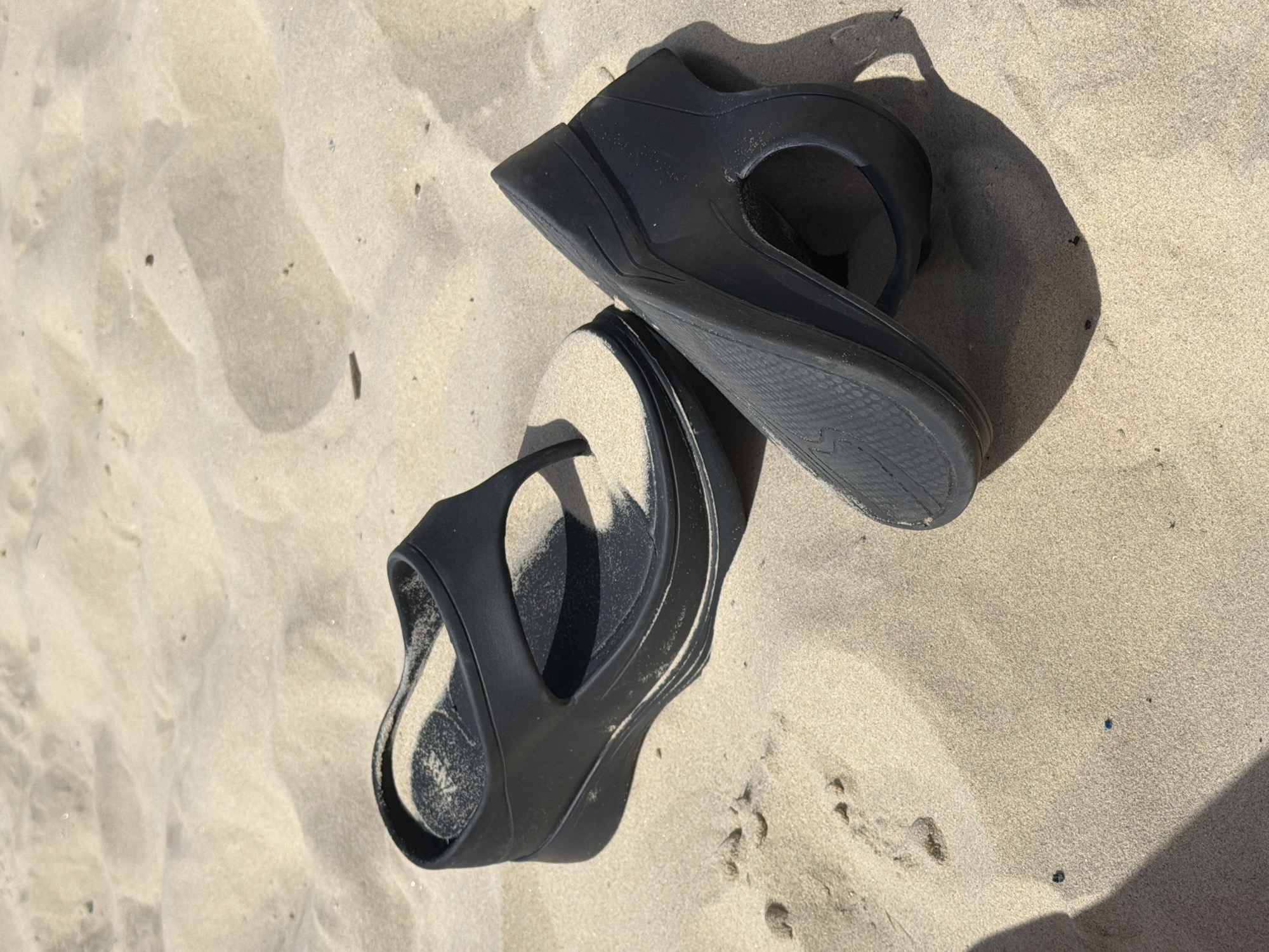 The image shows a pair of black flip-flop sandals lying on tan sand. Sand is inside of both sandals. A footprint is visible in the sand near the sandals. The location is likely a beach, potentially in Tel Aviv, Israel, based on the provided context. There are no people visible.
