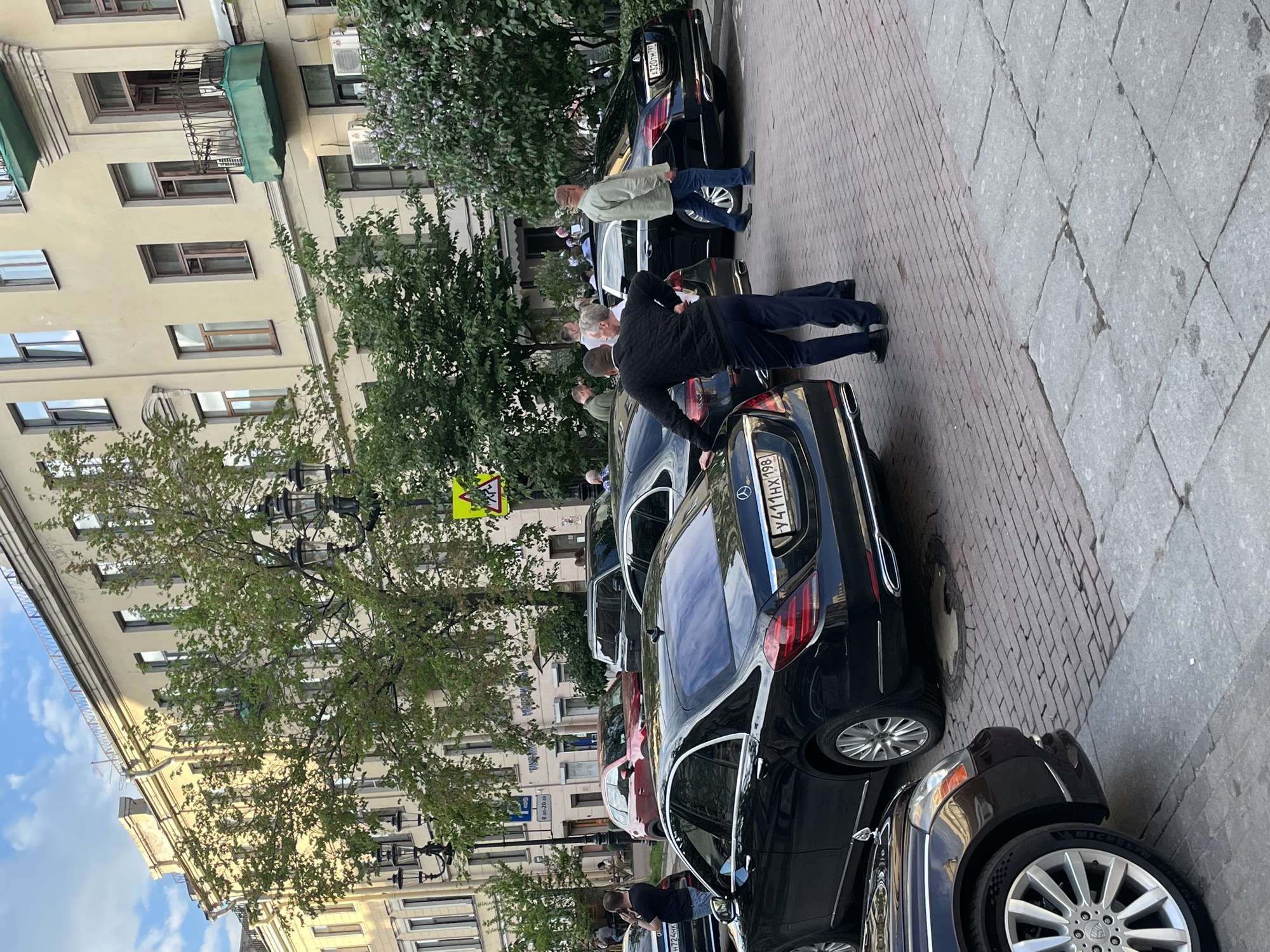 The image depicts a daytime street scene in what appears to be Saint Petersburg, Russia, with an emphasis on parked cars and people interacting around them. The sky is bright with some clouds, suggesting fair weather.
In the foreground, several vehicles are parked parallel to a multi-story building. A prominent black Mercedes-Benz sedan has its rear facing the viewer, displaying the Russian license plate "Y411HX 197." Another black car, partially visible behind it, bears the license plate "A320TM 97." A dark-colored Bentley is also partially visible in the lower right, with a portion of its license plate "H724HK" discernable.
Several men are present among the cars. One man, wearing a black quilted jacket and dark trousers, is leaning into the rear of the Mercedes-Benz, suggesting he is accessing its trunk. Another man in a light-colored jacket and jeans stands nearby. Other individuals are scattered further down the street, some gathered near the vehicles, others walking along the sidewalk.
The street features a distinctive pavement combining larger light-colored diamond-patterned tiles with a section of smaller, reddish-brown brick-like pavers. The background consists of multi-story buildings with numerous windows, some featuring balconies with green coverings and air conditioning units. Lush green trees line the street, adding natural elements to the urban setting.
Visible street signs include a yellow triangular warning sign with a black symbol and a blue rectangular sign that appears to indicate parking information, with "8:00 - 20:00" visible. The overall scene captures a slice of daily life in a European city, with a focus on street-side activity and parked vehicles.