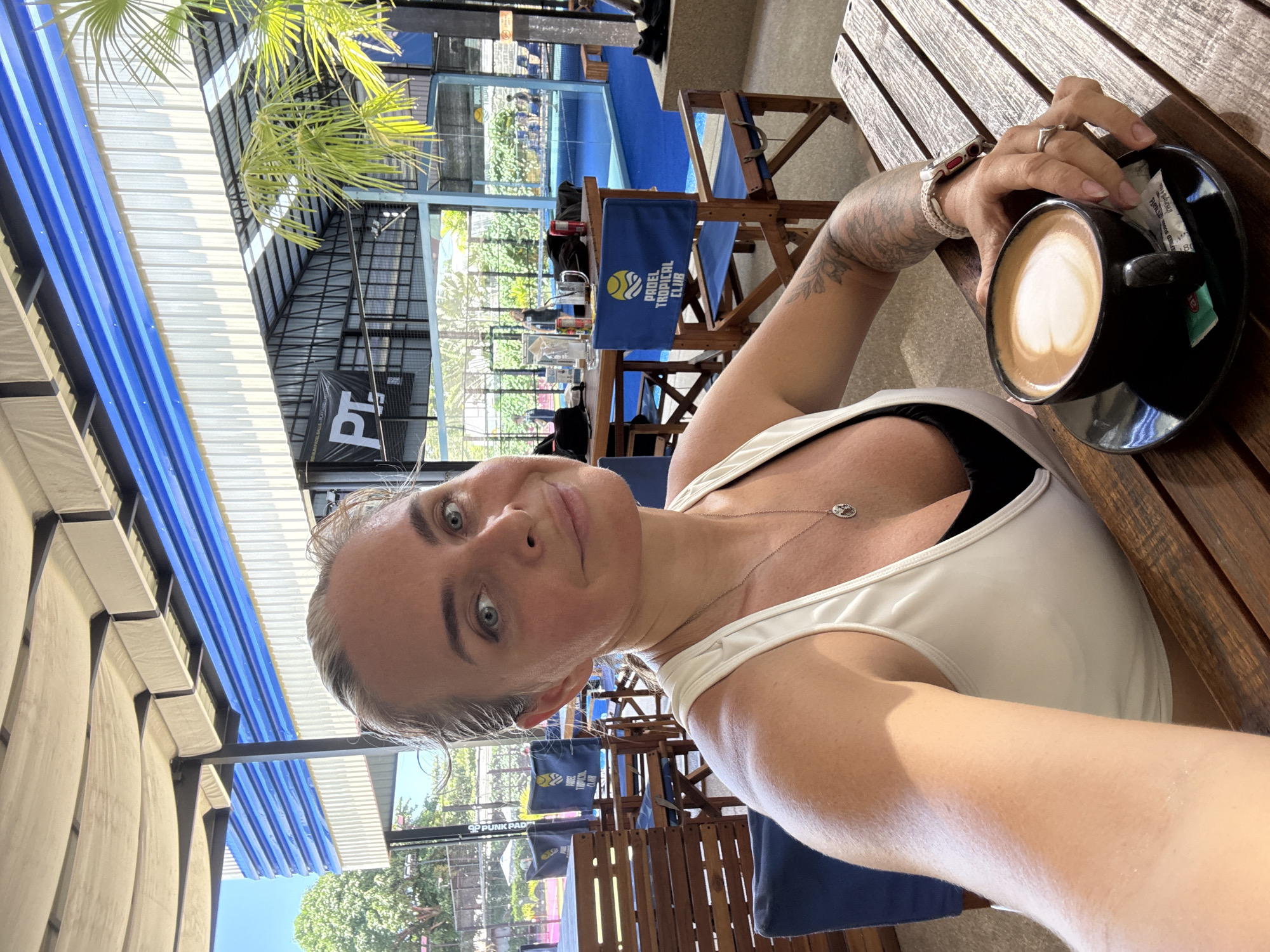 A woman with blonde hair pulled back, wearing a white tank top over a black sports bra, takes a selfie at an outdoor cafe at the Padel Tropical Club in Ban Bang Rak, Thailand. She has light blue eyes, is smiling slightly, and wears a silver pendant necklace. Her left arm, adorned with tattoos, rests on a wooden table where a black cup of coffee with latte art sits on a saucer next to a sugar packet. A wooden chair with a blue cushion bearing the club's logo is visible behind her. The background shows a covered patio with blue and white striped siding, palm trees, and glimpses of a blue padel court with players.