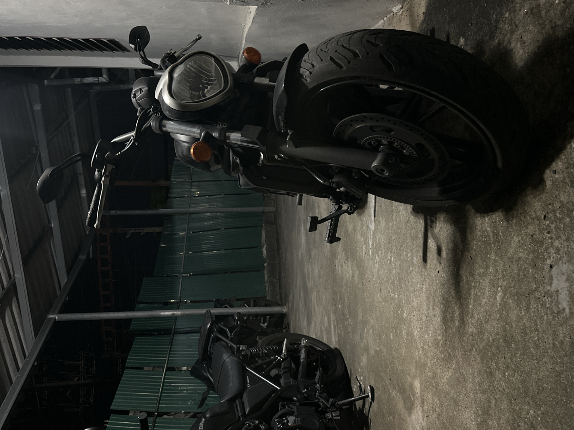 The image depicts two motorcycles parked indoors, possibly in a garage or workshop, with a concrete floor and a corrugated metal wall in the background. The lighting suggests it is nighttime or the area is dimly lit.
The motorcycle in the foreground is a black cruiser-style bike with a prominent headlight and handlebars. Its front wheel and part of the engine are visible. The rear wheel, with its tire tread and brake disc, is a focal point of the image, positioned towards the right.
Behind the first motorcycle, another black motorcycle is partially visible. Its seat, rear suspension, and part of its exhaust system can be seen.
The setting appears to be utilitarian and somewhat rough, with exposed structural elements and a slightly grimy concrete floor. The corrugated metal wall has a greenish hue, possibly due to lighting or the material itself. There are no people visible in the image. The overall mood is quiet and still, consistent with a parking area at night.
No visible text is clearly discernible in the image. The context provided indicates the location is Karon, Thailand.