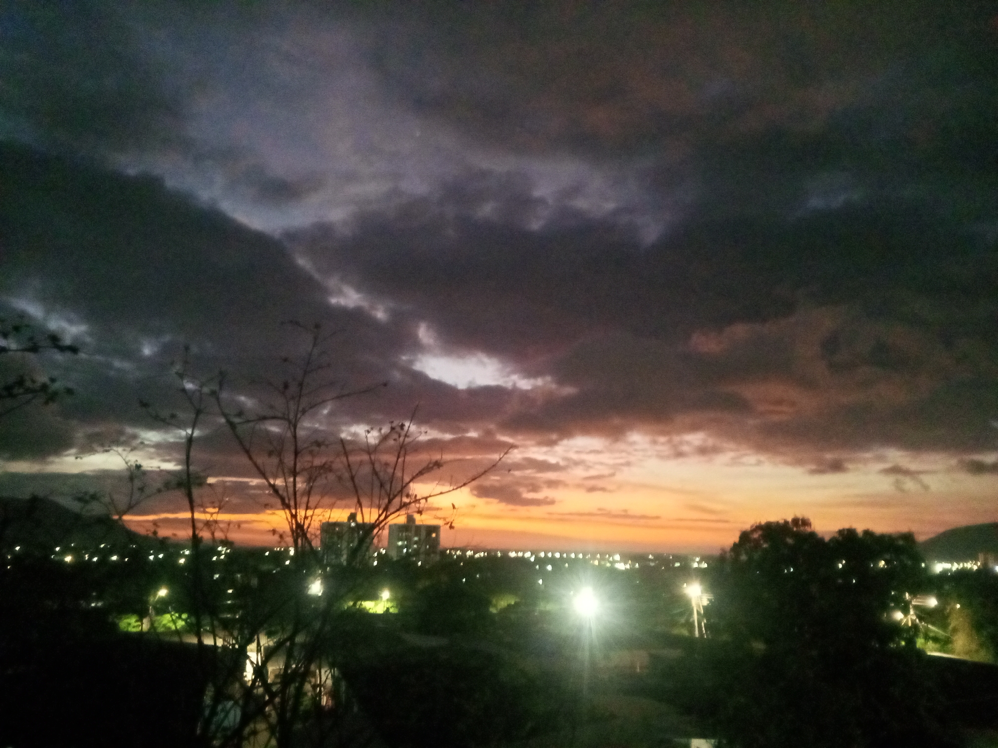 The image depicts a night scene in an urban environment. A sky filled with dark, layered clouds is visible, with the setting sun casting orange and purple hues along the horizon. In the foreground, bare tree branches are silhouetted against the twilight sky. Below the horizon, the lights of a city or town twinkle, illuminating streets and buildings. A cluster of larger buildings is discernible in the middle distance. Green foliage indicates the presence of trees and possibly parks. The location is Unknown, Unknown.