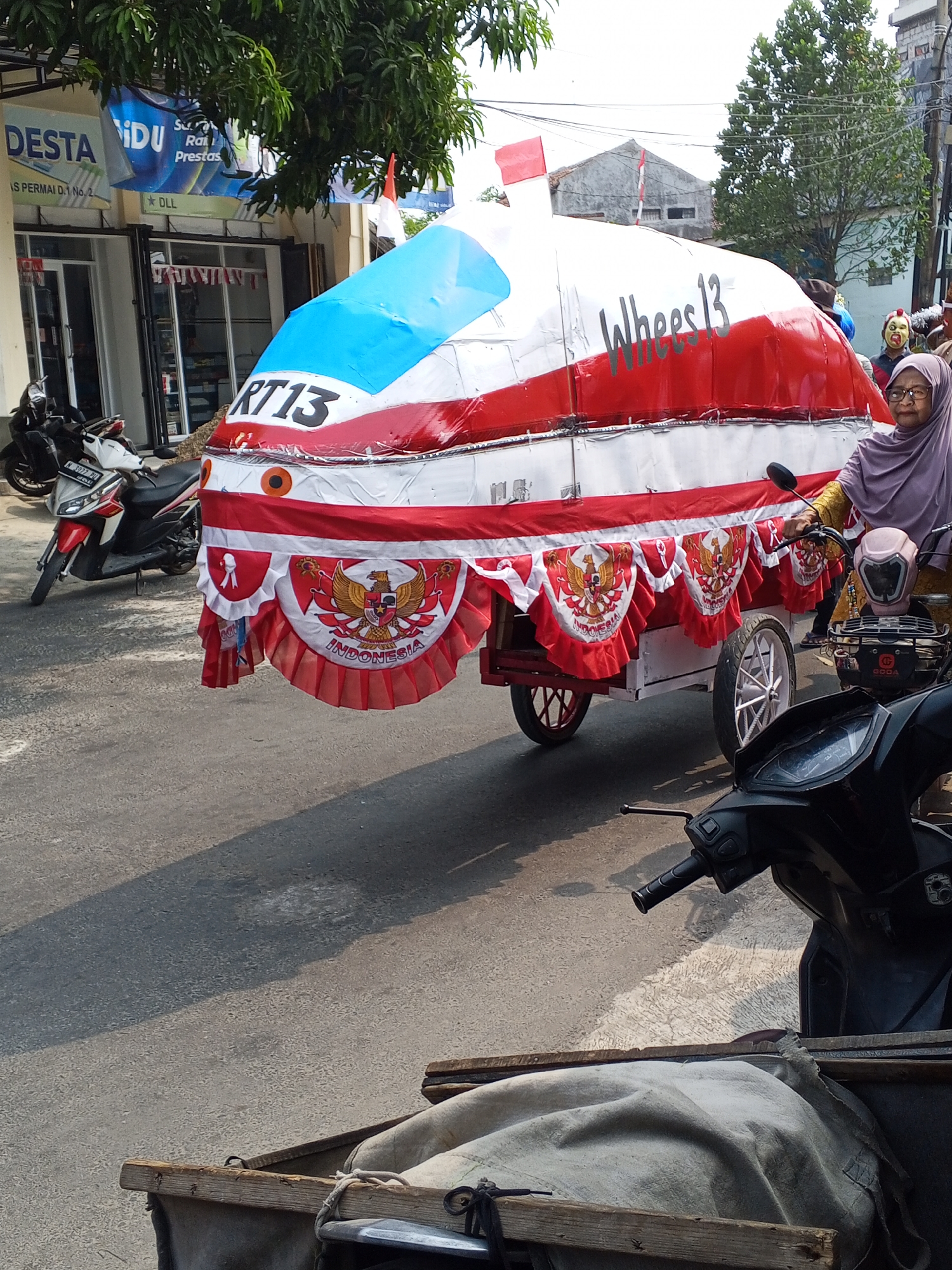 In Ciruas, Indonesia, a parade float shaped like a large capsule with red and white decorations is being pulled by a motorcycle. The float is adorned with the Indonesian coat of arms and the text "RT13" and "Wheels13". A woman wearing a hijab and a yellow top is operating the motorcycle. Another motorcycle is parked nearby. The scene takes place on a paved street with buildings and trees in the background. The atmosphere suggests a local event or celebration.