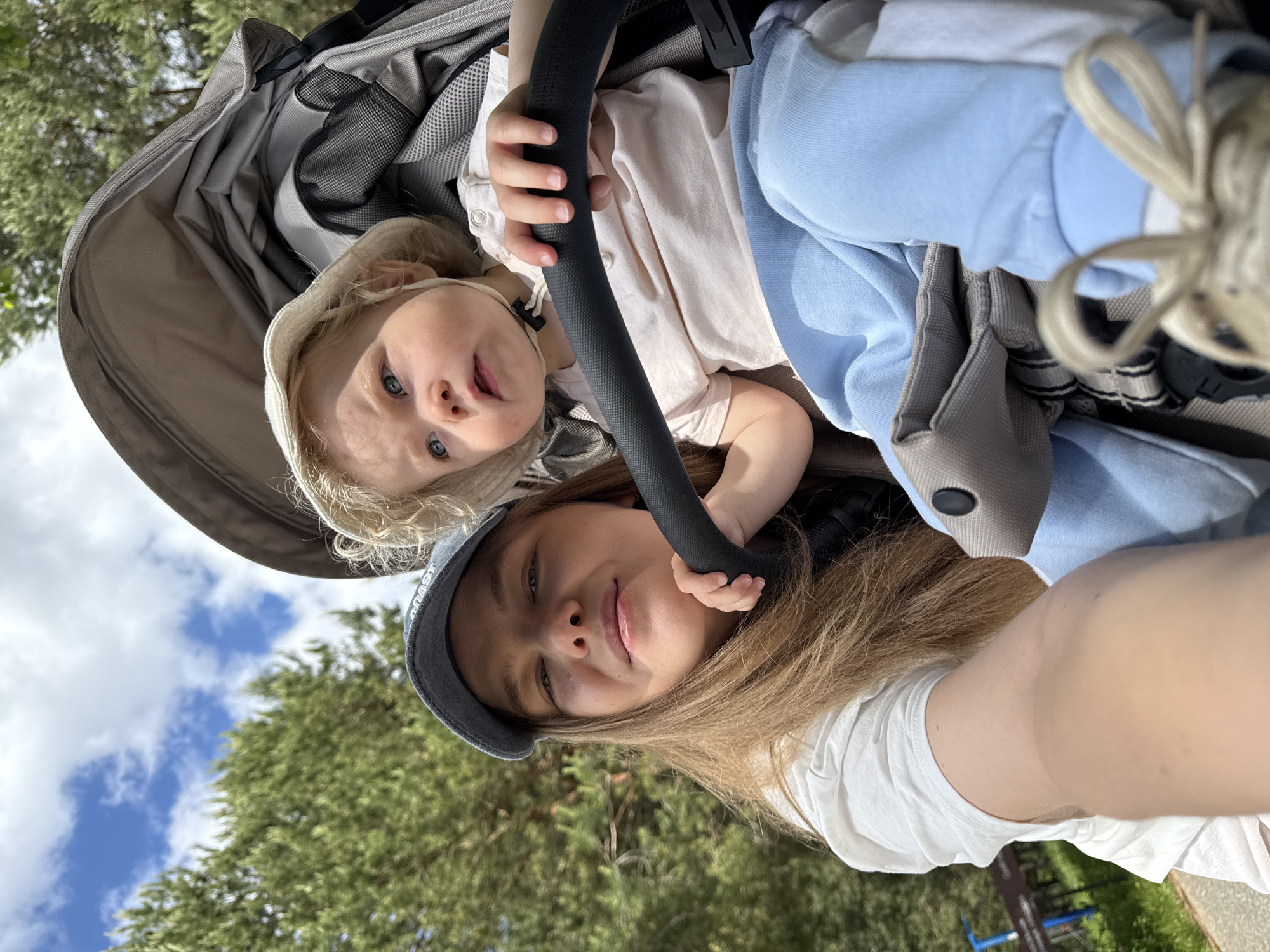 A bright, outdoor selfie features a young woman and a small child in Dobrograd, Russia. The image is composed from a low angle, looking upwards towards the subjects and the sky.
In the foreground, a young woman with long, light brown hair wears a white t-shirt and a dark grey baseball cap. The word "COAST" is visible on the brim of her cap. She has a calm expression, looking directly at the camera. Above her, a child, likely a toddler, is seated in a grey stroller. The child has blonde, curly hair, blue eyes, and is wearing a light-colored top, light blue pants, and a light hat. The child holds onto the stroller's black bumper bar and looks towards the camera with a curious, alert expression.
The scene is set outdoors amidst nature. Lush green trees form the middle ground, reaching up towards a bright blue sky dotted with scattered white clouds. The clear visibility and strong lighting indicate it is daytime with sunny weather. The overall activity suggests a relaxed outing, possibly in a park or similar green space.