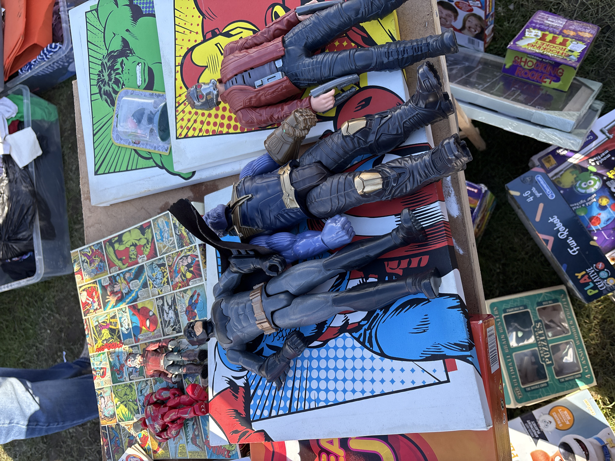 The image shows a variety of objects spread out on a possibly uneven surface, possibly grass or pavement. There are action figures of Batman, Thanos, and Star-Lord. They are placed on comic book-themed canvas prints or boards, including depictions of Hulk and Spider-Man. Other items present include board games, a "Shocking Rocket" prank toy, and a box of "Puzzles" with brain teaser. In the lower left corner, a person wearing blue jeans is partially visible.
The image has no specific information that confirms if it was taken in the city of Chirk, United Kingdom.