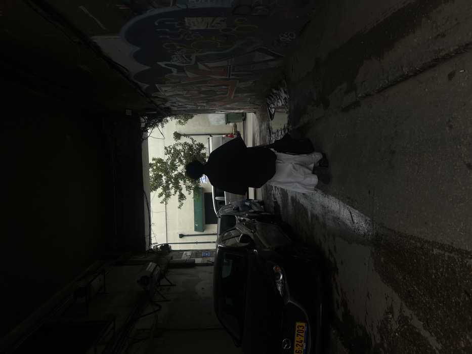 The image shows a dimly lit urban passageway in Tel-Aviv, Israel, viewed from within, looking towards a brighter exit. A single person, seen from behind, walks away from the viewer towards the illuminated outdoor area. The individual wears a dark, loose-fitting upper garment over a white, floor-length skirt or wide trousers, and carries a dark bag in their left hand.
The ground of the passageway is asphalt or concrete, visibly wet with standing water and tire tracks. On the left side of the passage, a dark-colored Mazda sedan is parked, displaying an Israeli license plate "624-703". Visible infrastructure on the left wall includes conduits and pipes. The right wall of the passage is extensively covered with multi-colored graffiti, featuring various tags and some discernible Hebrew script. Exposed wiring and cables are present overhead near the exit.
Beyond the passageway's opening, a light-colored building with a green awning is visible, alongside green foliage from a tree and a green refuse bin positioned to the right.