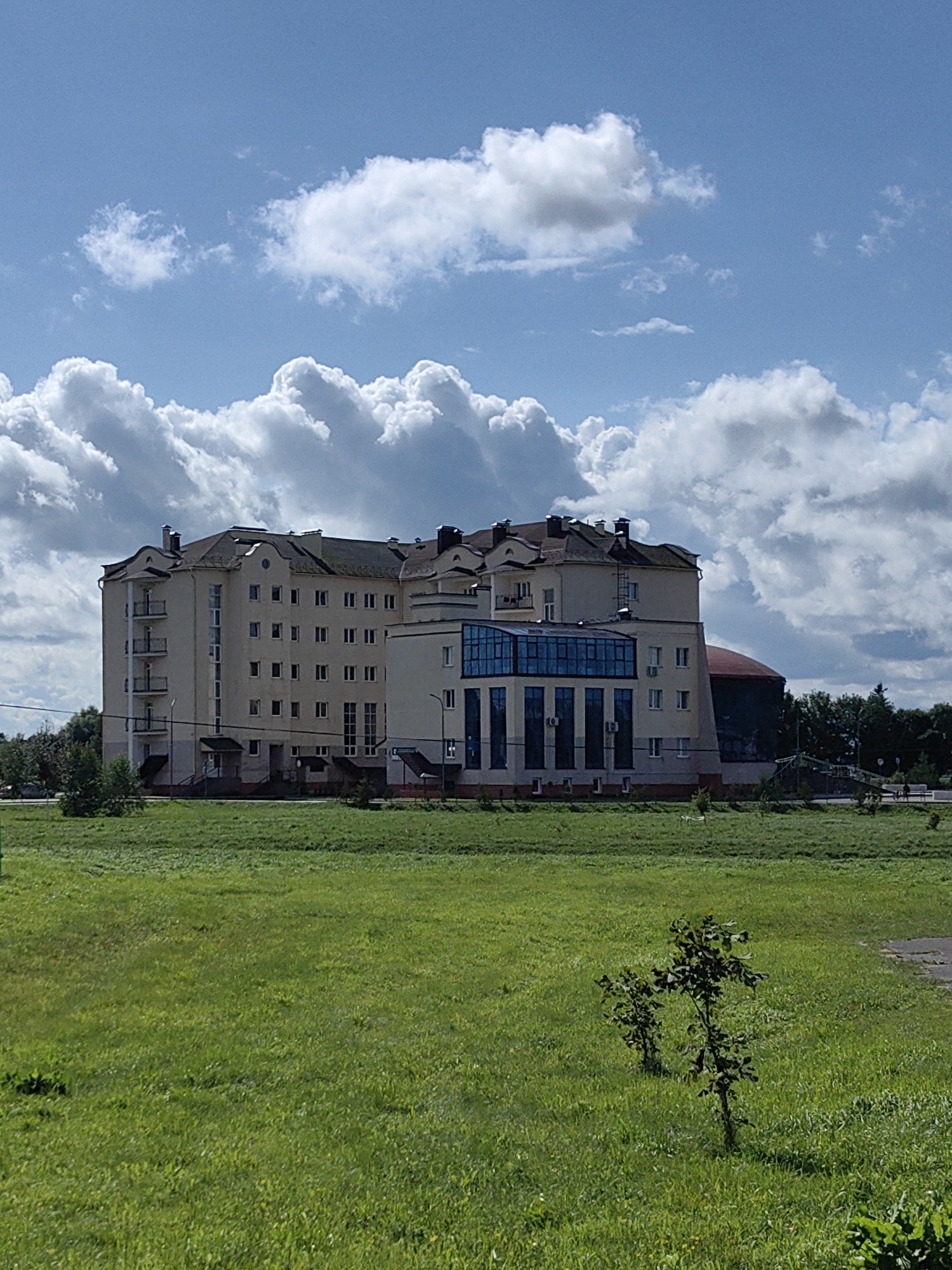 The image captures a multi-story beige building with a dark-colored roof situated in Myadzyel, Belarus. The building features numerous windows, balconies on the left side, and a modern section with large blue glass windows on the front. The building is surrounded by a vast expanse of green grass, with a few small trees visible in the foreground and a line of trees in the background. The sky above is a clear blue with large, fluffy white clouds. The scene appears to be a bright, sunny day.
