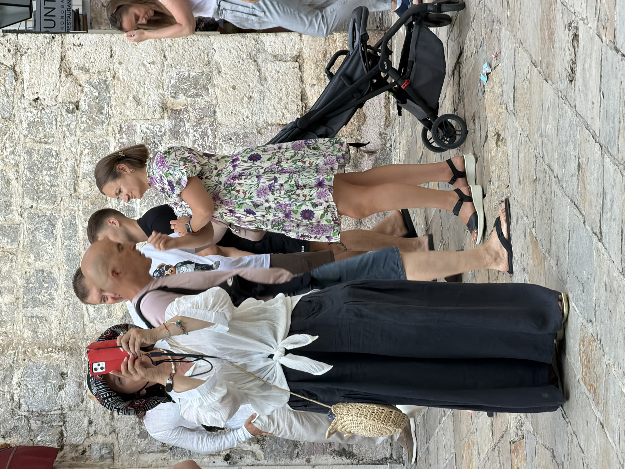 The image shows a group of people on a cobblestone street in Budva, Montenegro. The street is lined with old stone buildings, and there is a sign for a restaurant visible in the background. In the foreground, a woman is taking a picture with her phone, wearing a white blouse and a black skirt. To her right, a man is looking at her, and further to the right, a woman in a floral dress is walking with a stroller. Several other people are visible in the background, some walking and some standing. The scene appears to be a busy tourist area on a sunny day.