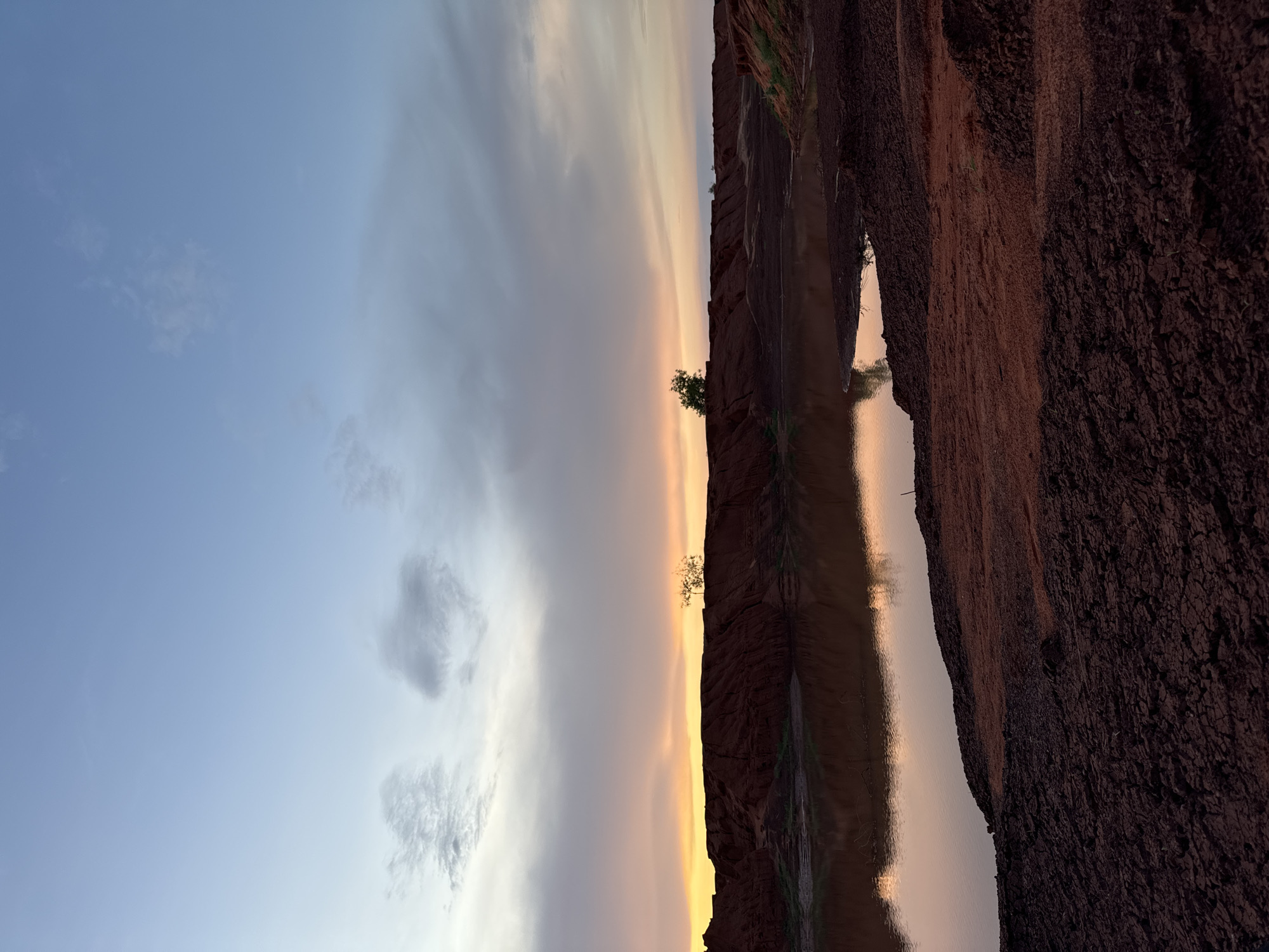The image, when correctly oriented, displays a serene natural landscape at either sunrise or sunset, indicative of a location like Mui Ne, Vietnam. A tranquil body of water, possibly a stream or narrow river, extends horizontally across the middle of the frame, serving as a reflective surface.
The sky, occupying the upper half, exhibits a captivating gradient of colors. It transitions from a clear, soft blue on the left to warm yellows, oranges, and subtle pinks towards the right and the horizon line. Sparse, wispy white and grey clouds are scattered across the sky, adding texture to its expanse.
On either side of the calm water are steep, textured banks or cliffs composed of reddish-brown earth and rock. The bank on the right, in the foreground, is significantly wider and closer, revealing intricate details of its rough, cracked, and dark surface. The bank on the left, across the water, appears as a thinner, darker silhouette against the colorful sky. A small, sparse tree with green foliage stands prominently on this distant bank, subtly breaking the horizon line.
The still water perfectly mirrors the sky's warm, glowing hues, creating a symmetrical reflection that intensifies the overall sense of color and light. The scene is devoid of any human activity, portraying a moment of profound natural beauty and tranquility. The soft, warm illumination strongly suggests either an early morning or late evening setting. The geological features, including the red earth and watercourse, align with the distinctive landscapes found in Mui Ne, Vietnam. No text is visible in the media file.