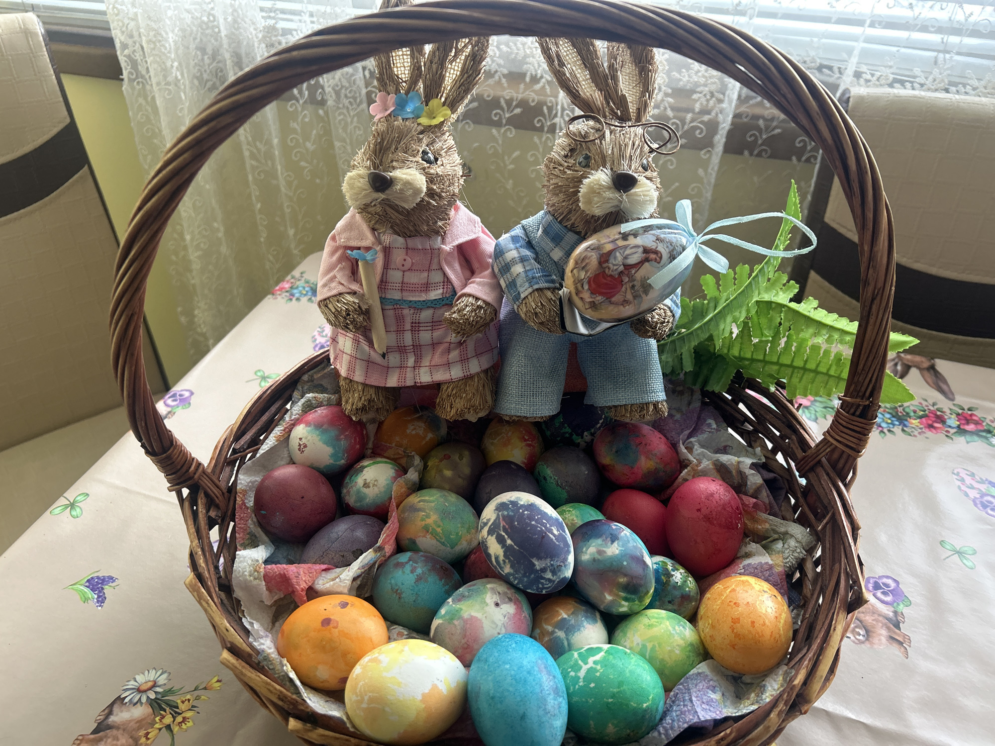 The image shows a woven wicker basket filled with dyed Easter eggs. Two straw-like bunny figurines stand amongst the eggs. The bunny on the left wears a pink plaid dress and has flowers in its hair. The bunny on the right wears a blue plaid shirt and eyeglasses, and is holding an egg decorated with a scene of children playing. Green fern fronds are visible near the right side of the basket. The basket sits on a tablecloth with a floral pattern. The location is Unknown, Unknown.
