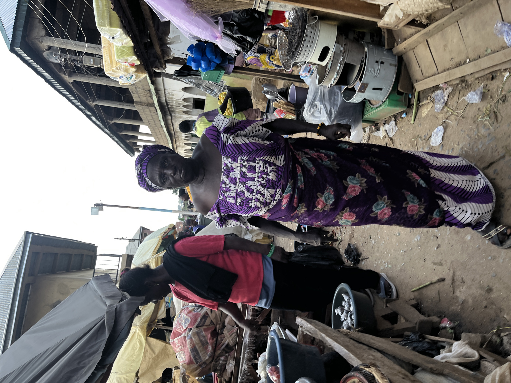 The image depicts an outdoor market scene in Rafin Par, Nigeria. In the foreground, an elderly woman stands facing the camera, dressed in a purple and white patterned traditional outfit and a matching headscarf. Her gaze is direct and somewhat stern. To her left, another woman in a red shirt and black skirt is partially visible, seemingly tending to goods in a market stall. The market appears to be bustling with activity, with various stalls and goods visible in the background. The ground is dusty and uneven, typical of an outdoor market. Items such as plastic containers, bags of produce, and household items are displayed for sale. The architecture in the background suggests a mix of traditional and modern buildings, with corrugated metal roofs and concrete structures. The overall atmosphere is one of a busy, active marketplace in a developing urban area.