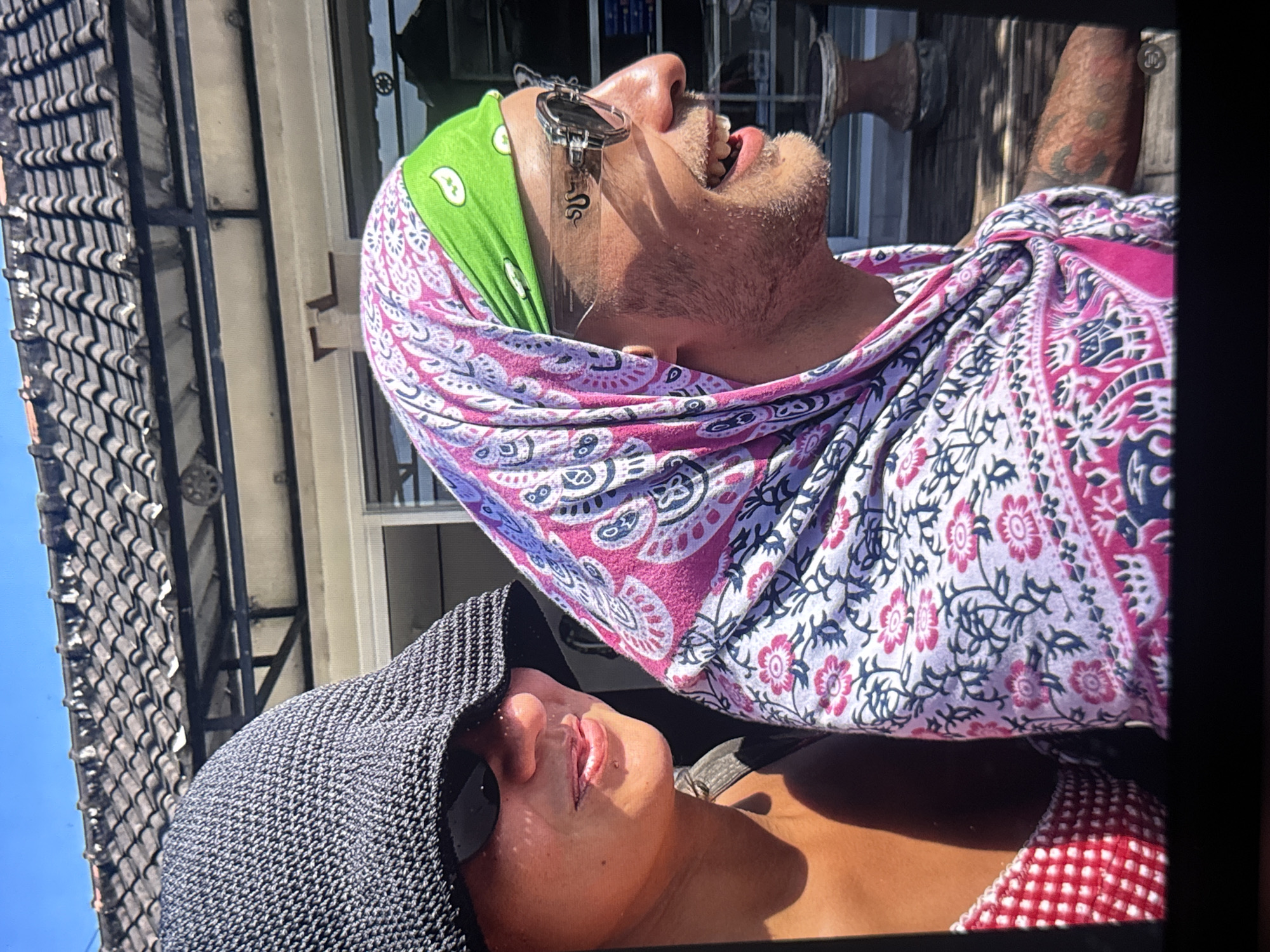 This media file depicts two individuals in an outdoor setting, possibly a residential area, given the tiled roof and building structures visible. The camera angle is unusual, with the image rotated, making it appear as if the subjects are lying down or are overhead.
The prominent subject, in the upper portion of the frame, is a person wearing a green bandana and a flowing garment with a pink, purple, and blue floral pattern. They have a beard and appear to be looking upwards with their mouth open. They are also wearing sunglasses with a dangling snake charm. The lighting suggests it is daytime, with bright sunlight casting shadows.
The second subject is partially visible in the lower portion of the frame, wearing a dark, textured hat and sunglasses. Their face is angled upwards, and their lips are parted. They are wearing a red and white checkered top. The overall mood seems relaxed and possibly candid. There is no visible text in the image. The location context is Kuta Selatan, Indonesia.
