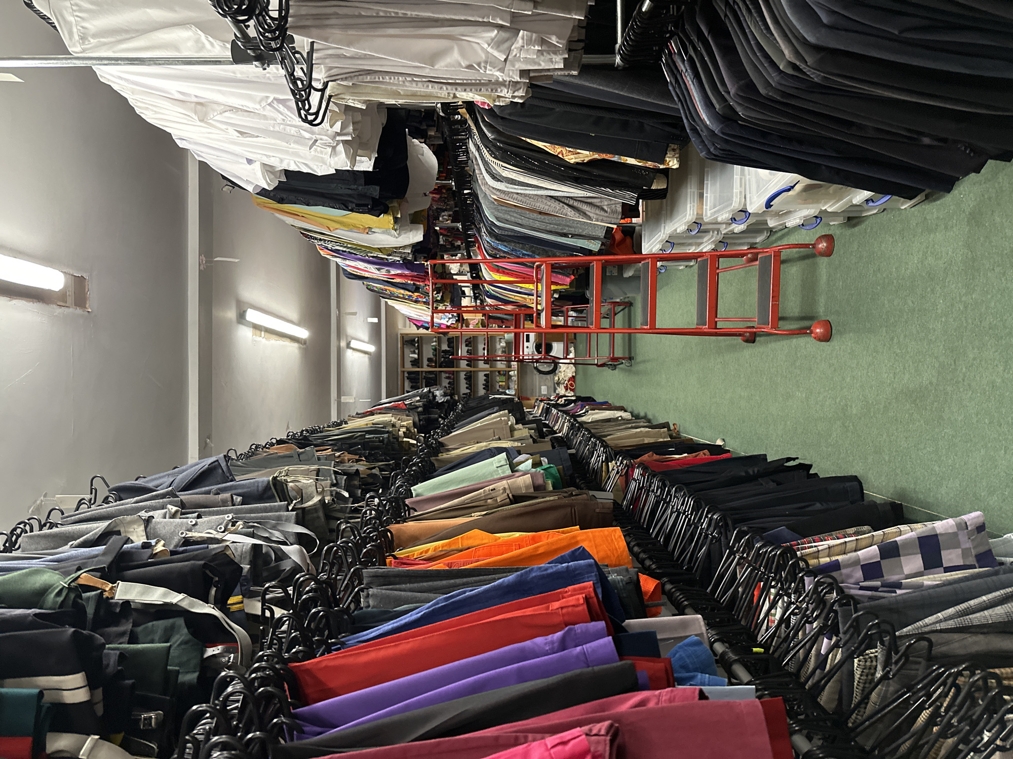 The image depicts a storage room filled with racks of clothing. The clothing appears to be sorted by type and color, with white shirts at one end, followed by other colored shirts, and trousers. A red step ladder sits on the green floor. The lighting is provided by fluorescent lights on the ceiling. There are no people visible in the image. The location is not determinable as London, United Kingdom.