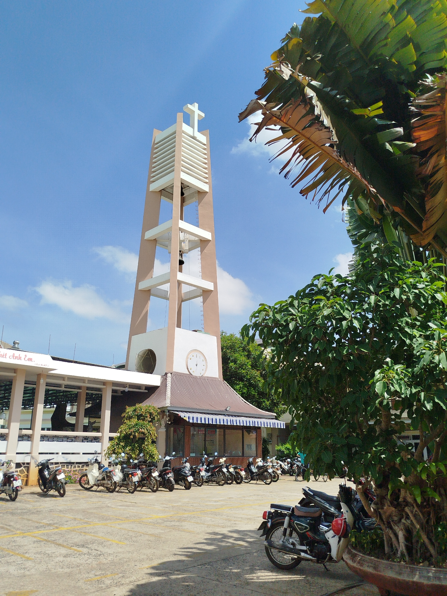 The image depicts a church bell tower in Pleiku Ward, Vietnam, under a partly cloudy sky. The beige and white tower is topped with a Christian cross. A clock face is visible on the tower's lower section. The base of the tower connects to a building with a brown roof and blue and white striped awning. Parked in front of the building are numerous motorbikes. Lush green foliage from trees and a large banana plant frame the right side of the image. A sign with Vietnamese script is visible on the building to the left.