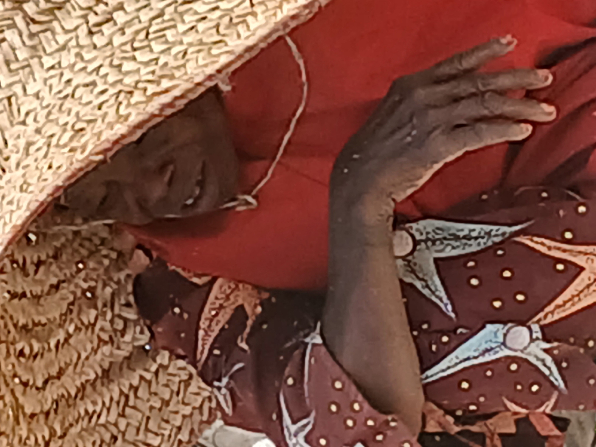 The image depicts a person with dark skin lying down under a woven straw hat. The person is wearing a red garment under a maroon and white patterned shirt or dress. One dark-skinned hand is visible. The location is Tudun Nupawa, Nigeria.