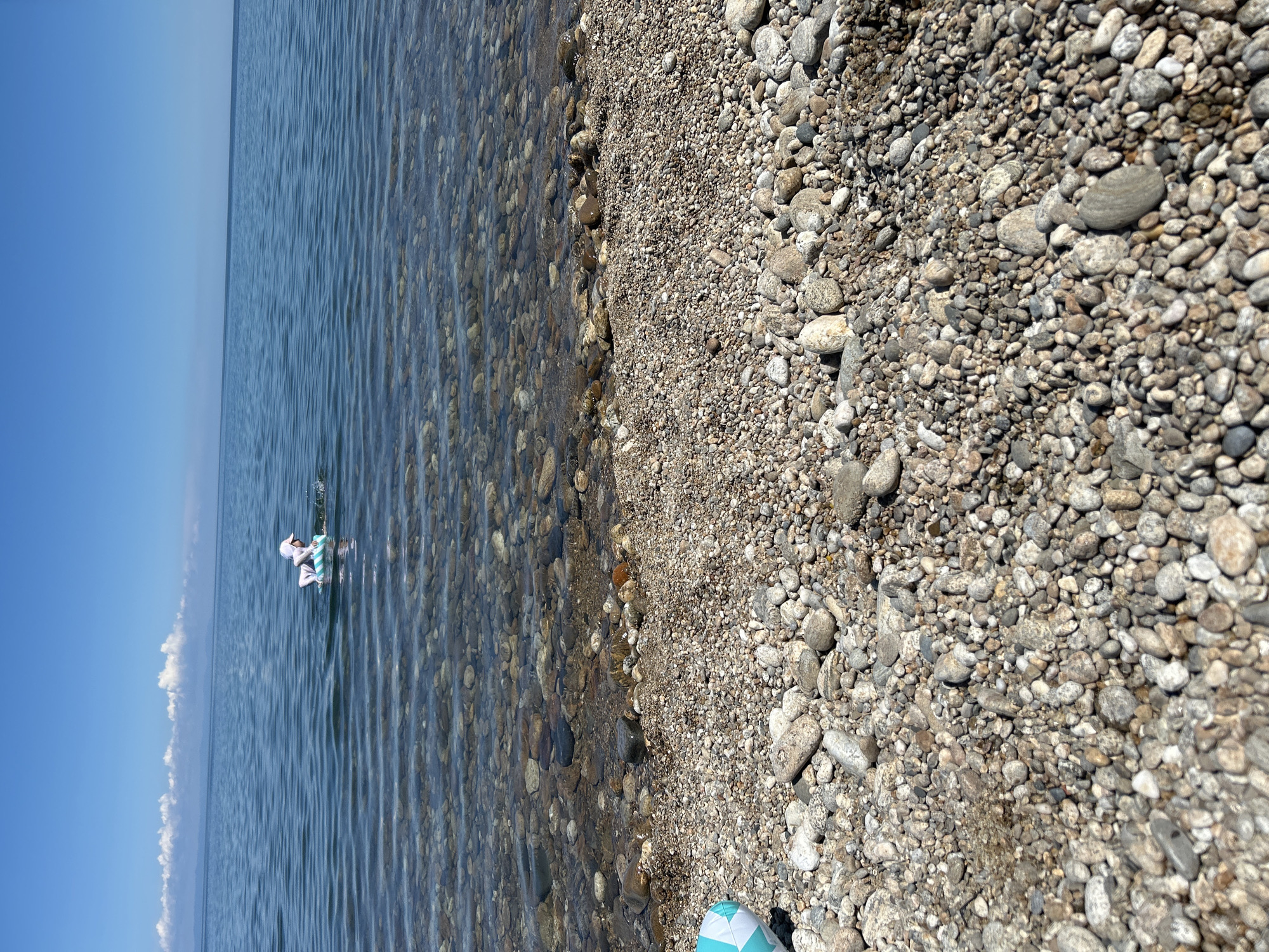 A person in a white hat and striped swimwear floats on a circular inflatable ring in clear, calm water. The shoreline, composed of small, smooth pebbles, slopes gently into the water. In the foreground, a portion of a blue and white patterned inflatable object rests on the pebbles. The sky above is a clear, bright blue with a few wispy white clouds on the horizon. The location is the shore of Lake Baikal in the village of Tolbazikha, Russia.