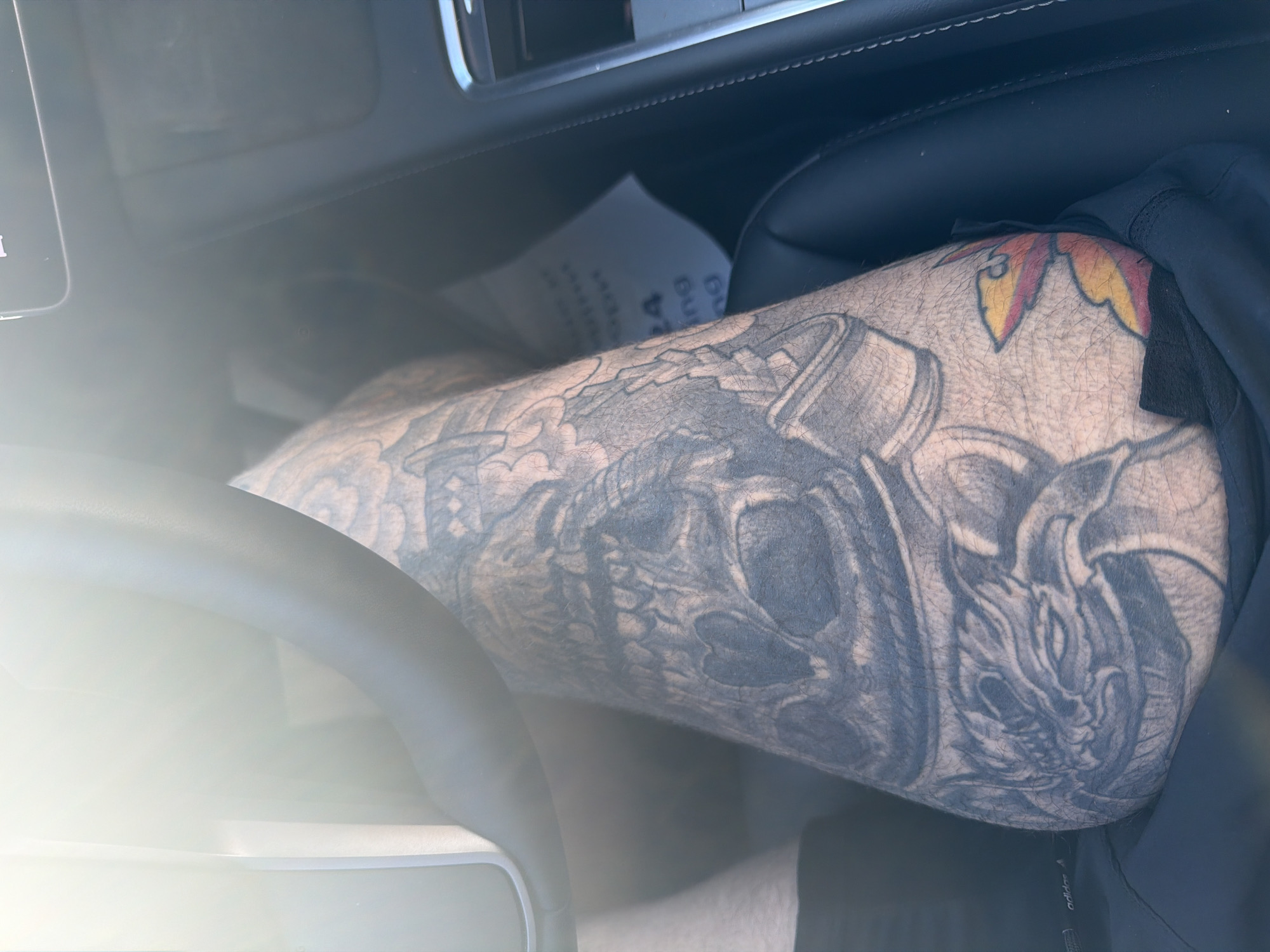 The image shows a close-up view of a person's leg, heavily tattooed with intricate designs, including a skull with a heart, a dragon's head, and a colorful abstract shape. The leg is positioned inside a vehicle, with parts of the steering wheel and car interior visible. The overall lighting suggests a bright, sunny day, with some lens flare present in the upper left portion of the image. The background is out of focus, but appears to be the interior of a car, possibly a modern one with a dark leather seat and dashboard. The leg is resting on what seems to be dark fabric, possibly clothing. The tattoo style is dark and detailed, with shading and line work.