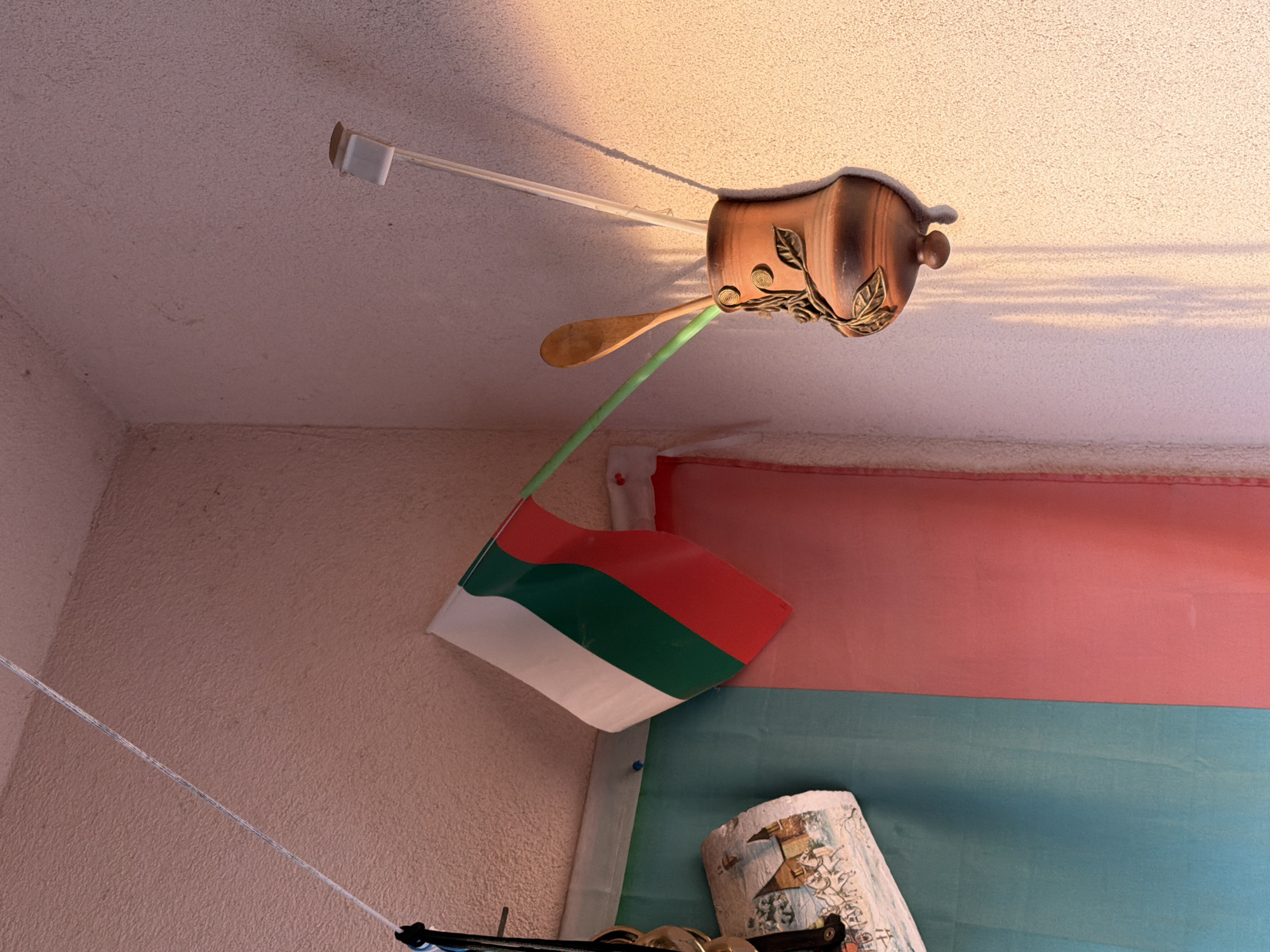 The image shows a room interior with a decorative light fixture attached to a textured ceiling. The light fixture is made of a dark brown material and has a vine-like design with leaves. Extending from the light fixture are a clear plastic rod and a wooden spoon. A green straw or tube connects the light fixture to a red, green, and white flag hanging on the wall. The wall is pinkish beige and slightly textured. A pink and blue striped fabric backdrop is behind the flag. A rolled-up piece of paper with a drawing is on the bottom right. A string extends from the bottom left and is attached to an object. The city and country are unknown.