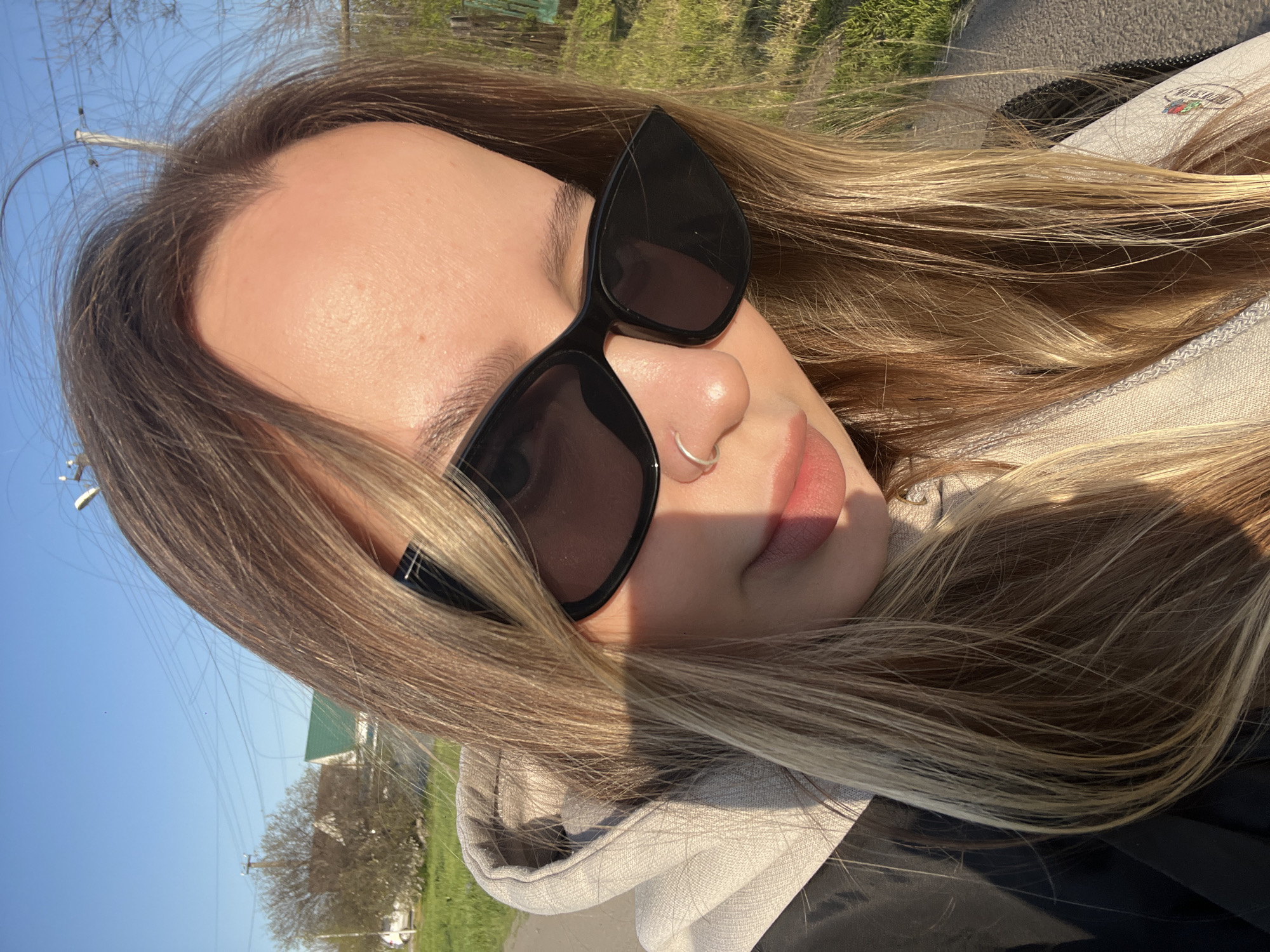 The image is a close-up, tilted portrait of a person, likely a woman, captured outdoors on a bright, sunny day. Her head is angled to the right, with her forehead, nose, lips, and part of her cheek visible. She has long, light brown hair with blonde highlights, which frames her face and extends around her.
She wears dark, cat-eye style sunglasses, obscuring her eyes. A small, thin silver hoop nose ring adorns her right nostril. Her lips are slightly parted, showing a natural pink hue. She is dressed in a light-colored (possibly beige or light grey) hooded sweatshirt, with the hood visible around her shoulders.
The sunlight creates distinct shadows across her face and clothing, indicating a high sun position. The background is out of focus but reveals a clear blue sky, green foliage, trees, and power lines, suggesting an outdoor, possibly rural or suburban setting. A distant building with a green roof is also faintly visible. On a tag or logo on her clothing, the text "FRUIT OF THE LOOM" is partially discernible. The overall scene conveys a relaxed, casual moment in pleasant weather.
