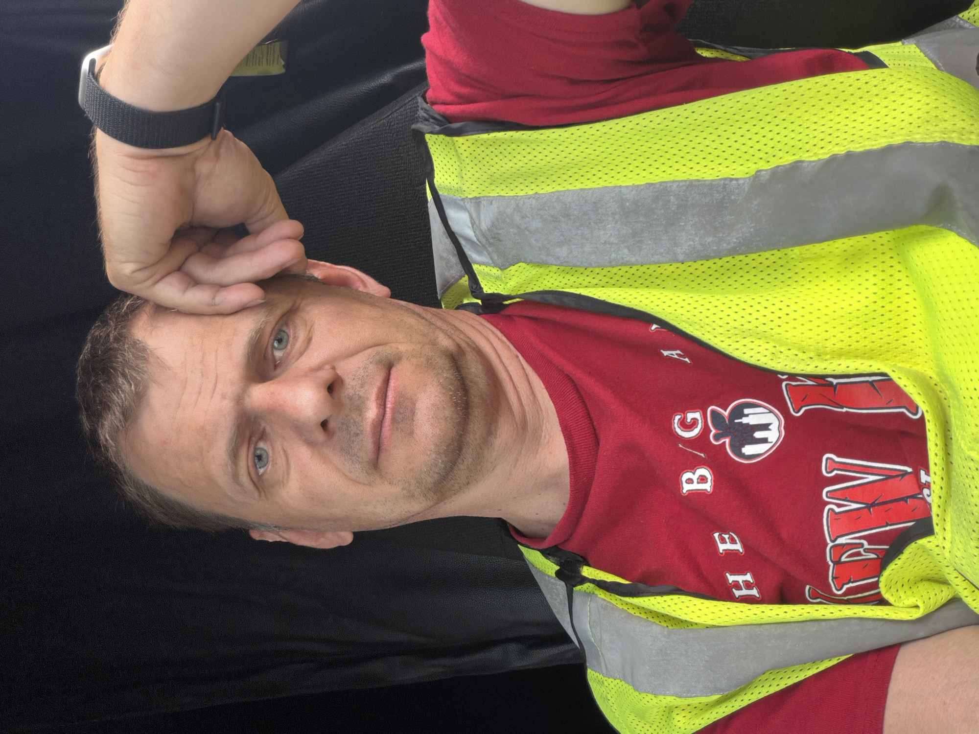 A man lies on his side, his head propped up by his hand. He is wearing a red t-shirt with white text and graphics, and a bright yellow safety vest with gray reflective stripes. The background is dark and appears to be an interior space, possibly a car seat. The man has light blue eyes and short, light brown hair. He has a slightly stubbled jawline and his expression is neutral. The lighting suggests it might be daytime, but the indoor setting makes it difficult to determine the exact time or weather. There is some visible text on his t-shirt, though it is partially obscured by the safety vest. Some legible letters include "H E B / G" and a partial word that appears to be "W V". There is also a graphic of a stylized building or landmark. The image is oriented sideways.