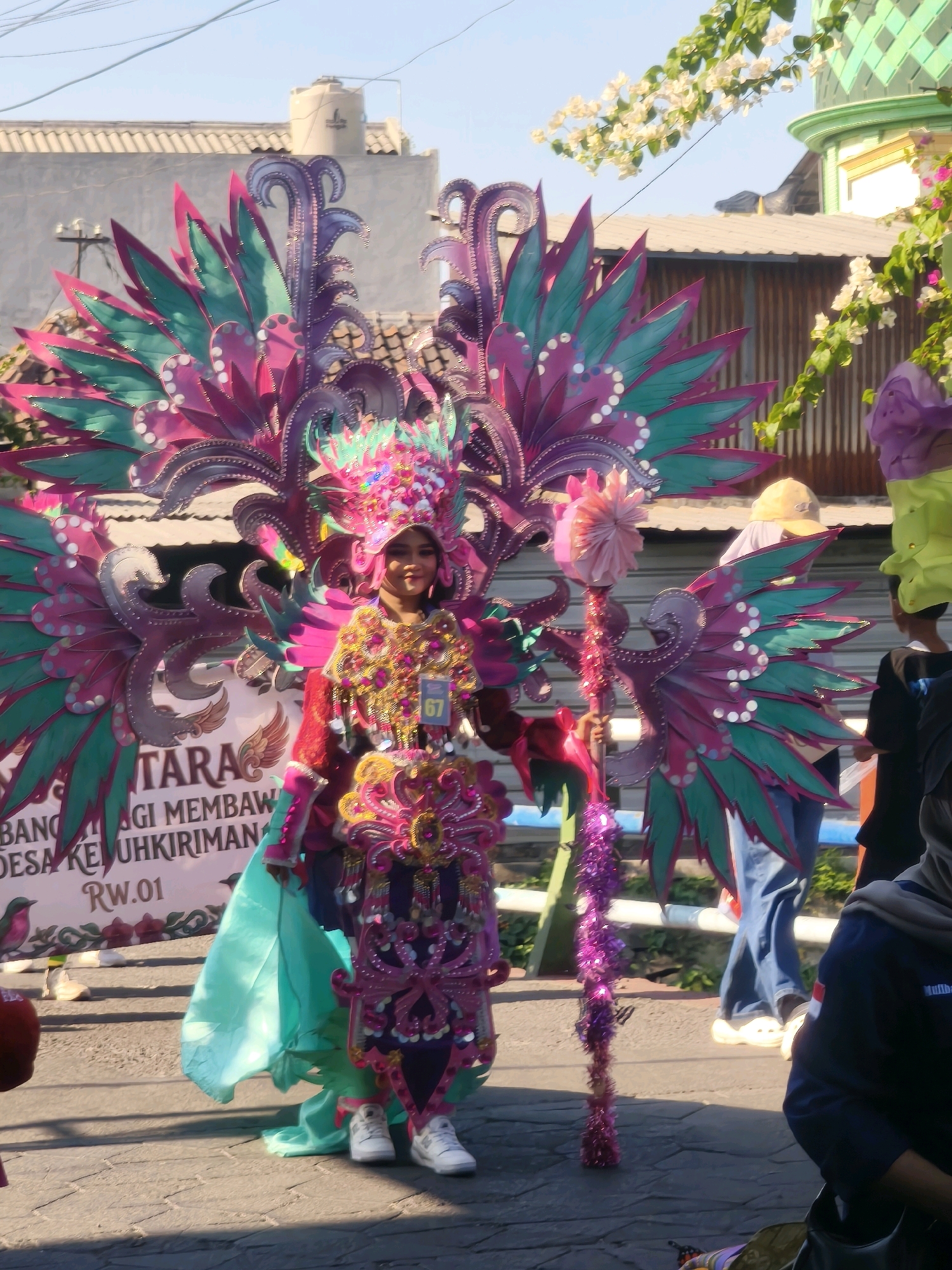 In Kepuh Kiriman, Indonesia, a young woman, adorned in an elaborate, colorful costume resembling butterfly wings and a headdress, stands on a paved street. The costume is predominantly purple, pink, and turquoise, embellished with sequins and other decorative elements. She holds a staff decorated with purple tinsel. A banner behind her, partially visible, reads "UNTARA BAGI MEMBAWA KESEUH KIRIMAN RW.01". The background features buildings with tiled roofs, a water tank, and blooming white flowers. Sunlight casts shadows, indicating it is daytime. Other individuals are present in the background, including one wearing a yellow hat and another in a dark shirt. The scene suggests a festive event or parade.