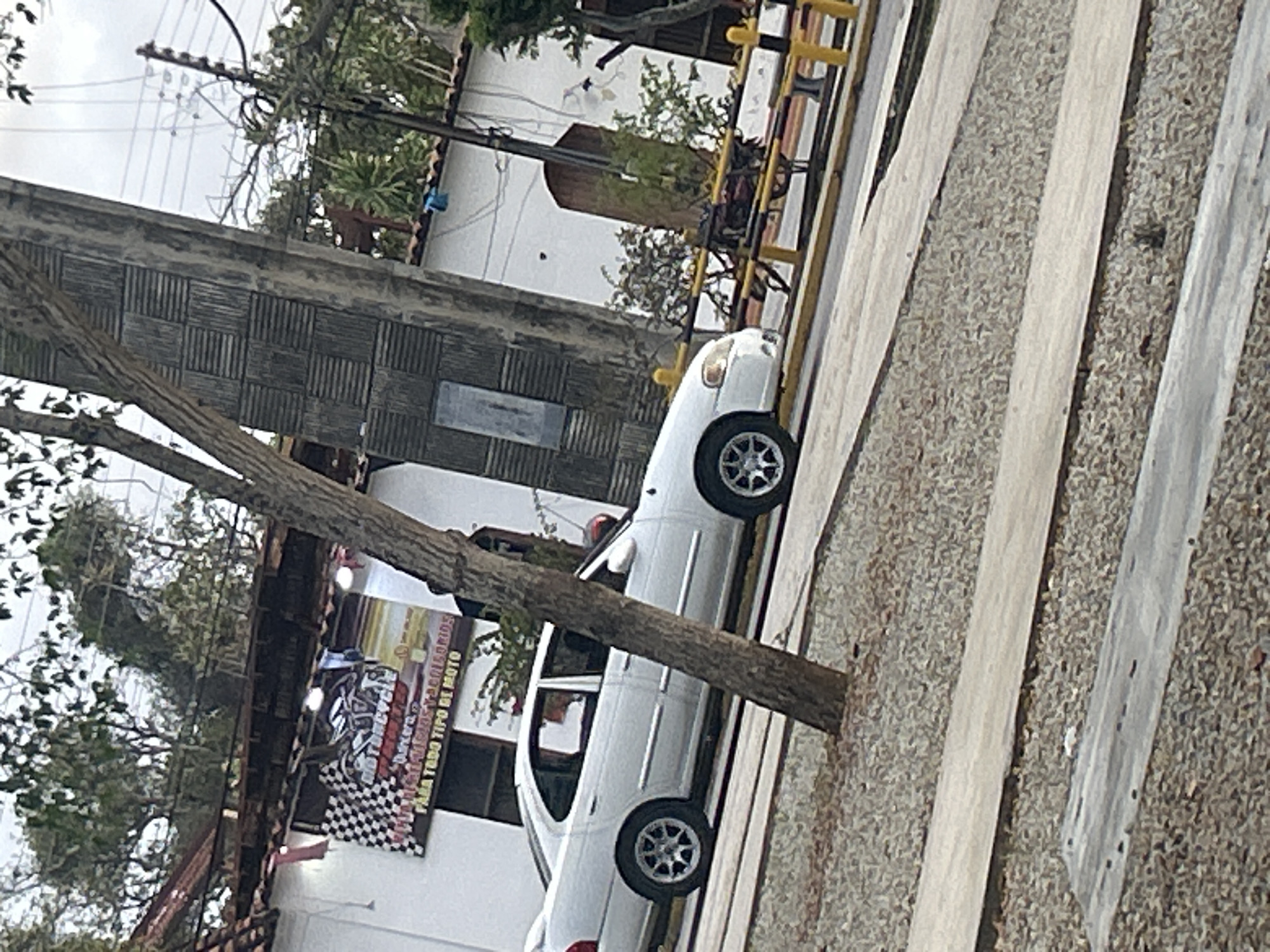 A white sedan is parked in front of a building in Santa Ana, Venezuela. The building has a white facade with a dark-tiled roof. A checkered flag banner hangs from the building. There are trees in the foreground and background, and a utility pole with wires is visible. The ground is covered in gravel and concrete.