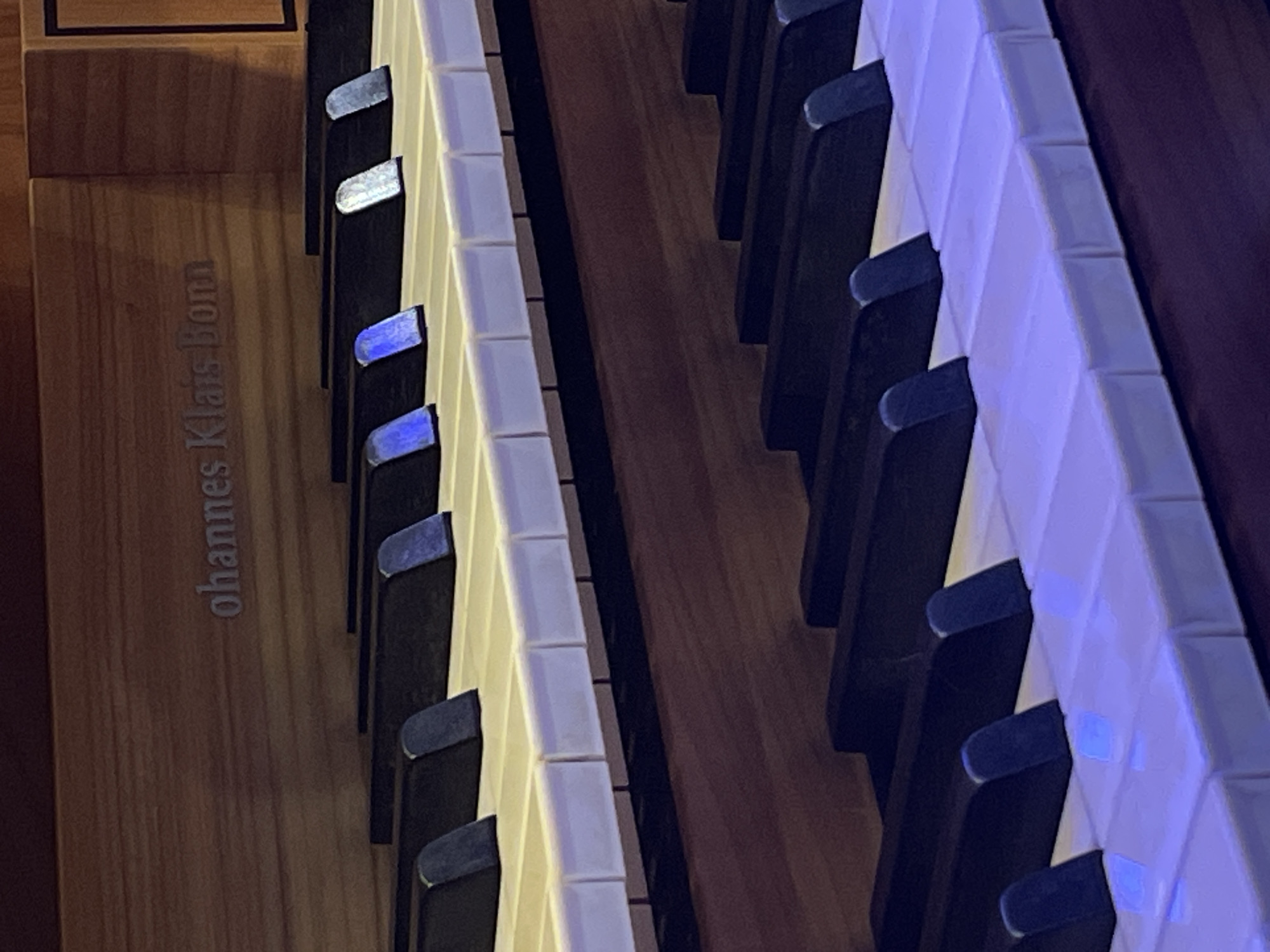 The image displays a close-up, angled view of two manuals (rows of keys) of a keyboard instrument, likely an organ or harpsichord. The perspective is from above and to the left, looking down the length of the keys. No people are visible.
The left manual features off-white to yellowish keys, with some appearing illuminated by a blue or purple light. Interspersed are dark, black keys, some with reflective, silvery-blue surfaces on their tops. The right manual also shows similar off-white/yellowish keys, many reflecting a distinct blue-purple light, along with dark keys. The keys on both manuals exhibit a segmented appearance, typical of organ or harpsichord keys.
To the left of the keys, a light-brown wooden panel runs vertically, part of the instrument's casing. Between the two manuals, there is a darker wooden panel. The lighting suggests an indoor setting, with artificial illumination creating vibrant blue and purple reflections on the keys.
Visible text engraved on the light-brown wooden panel to the left of the keys reads "Johannes Klais Bonn". This identifies the instrument as a creation by Klais Orgelbau, a renowned German organ builder based in Bonn. Although the instrument itself is of German origin, the location context provided for the image is Yekaterinburg, Russia, indicating where this particular instrument is situated. The image focuses solely on the intricate details of the instrument's keyboard, capturing its materials and the interplay of light.