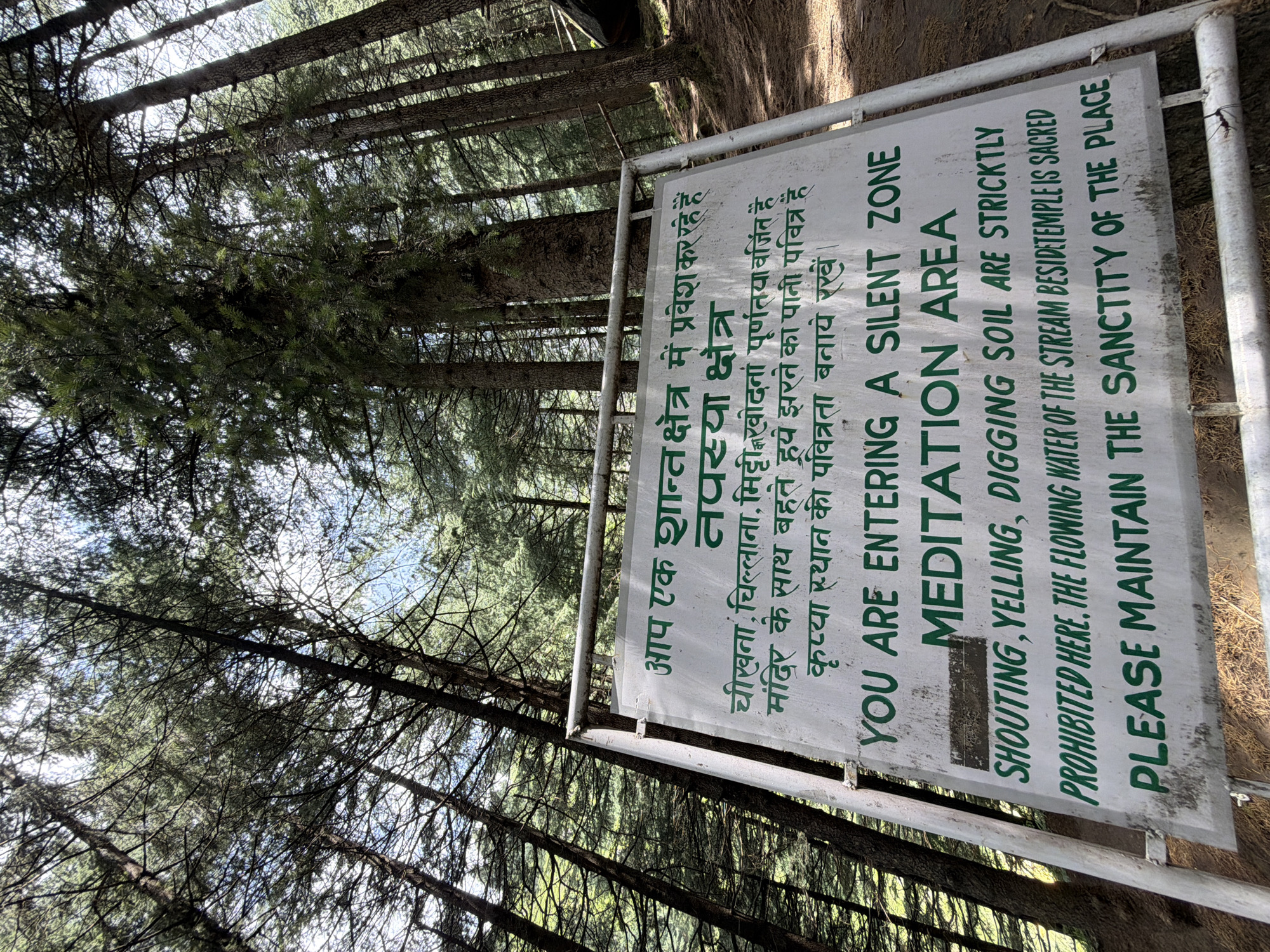 A white sign with green text is displayed in a forest setting, likely in Bahang, India, as indicated by the Hindi script on the sign. The sign is mounted on a metal frame and stands amidst tall trees with dense foliage. The text on the sign, in both Hindi and English, states that visitors are entering a silent zone and meditation area. It prohibits shouting, yelling, and digging soil, and emphasizes that the flowing water of the stream beside the temple is sacred. Visitors are requested to maintain the sanctity of the place. The ground around the sign appears to be covered with dry leaves and dirt. The sunlight filters through the trees, creating dappled light on the ground. There are no people or animals visible in the image.