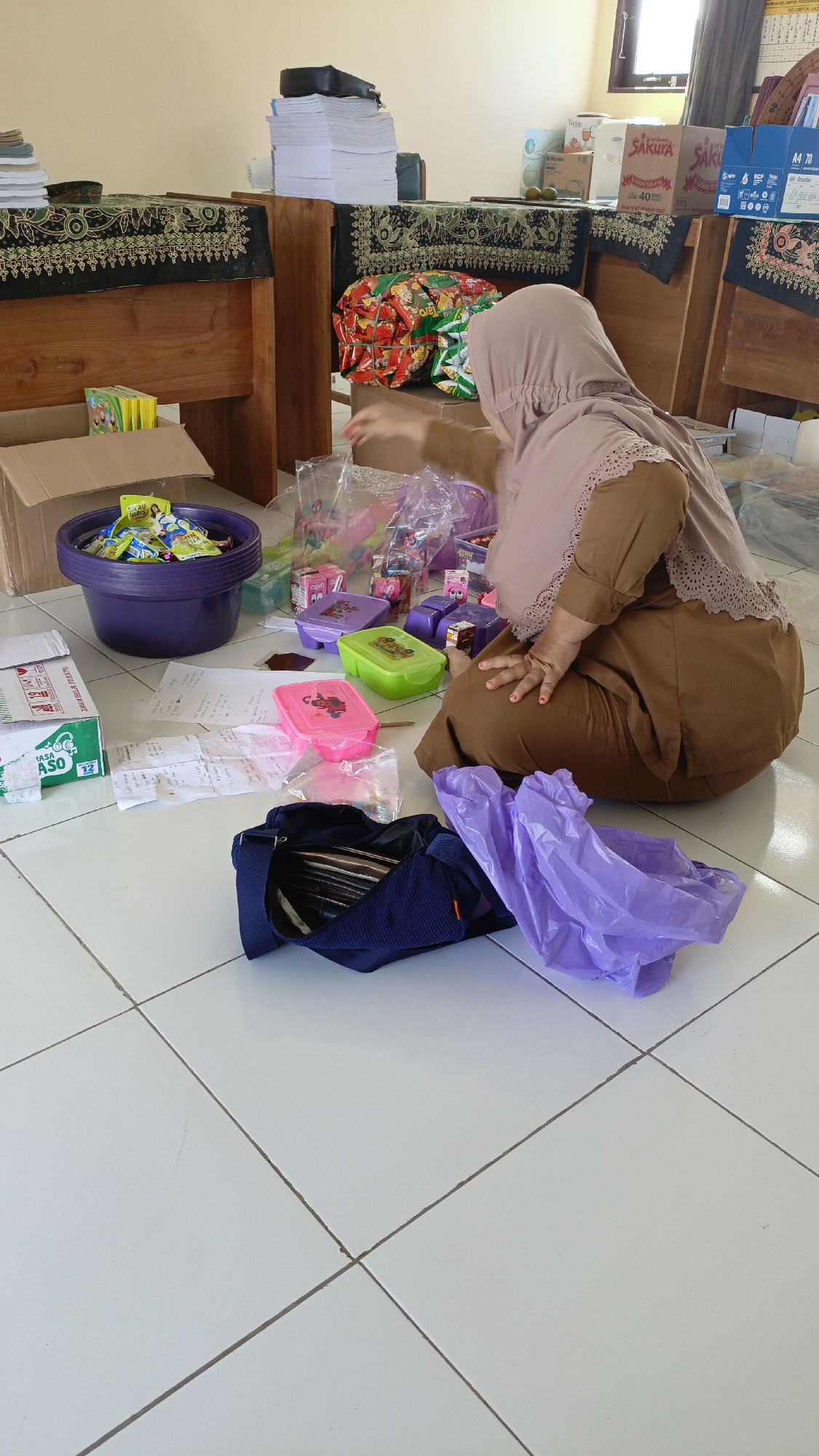 In Saring Sungai Binjai, Indonesia, a woman wearing a brown hijab and brown attire is kneeling on a tiled floor in what appears to be a classroom or office. She is sorting through a collection of small plastic containers, some of which are brightly colored pink and green, filled with small items, possibly candy or toys. A large purple plastic bowl filled with similar small items is to her left, situated next to an open cardboard box. Various other plastic bags and packages are scattered around her. Behind her, to the right, are several boxes, some labeled "SAKURA." Stacked papers are visible on wooden tables in the background, which are covered with patterned cloths. A navy blue bag lies open on the floor near her, and a crumpled purple plastic bag is next to it. The overall scene suggests the preparation of gift bags or distribution of items.