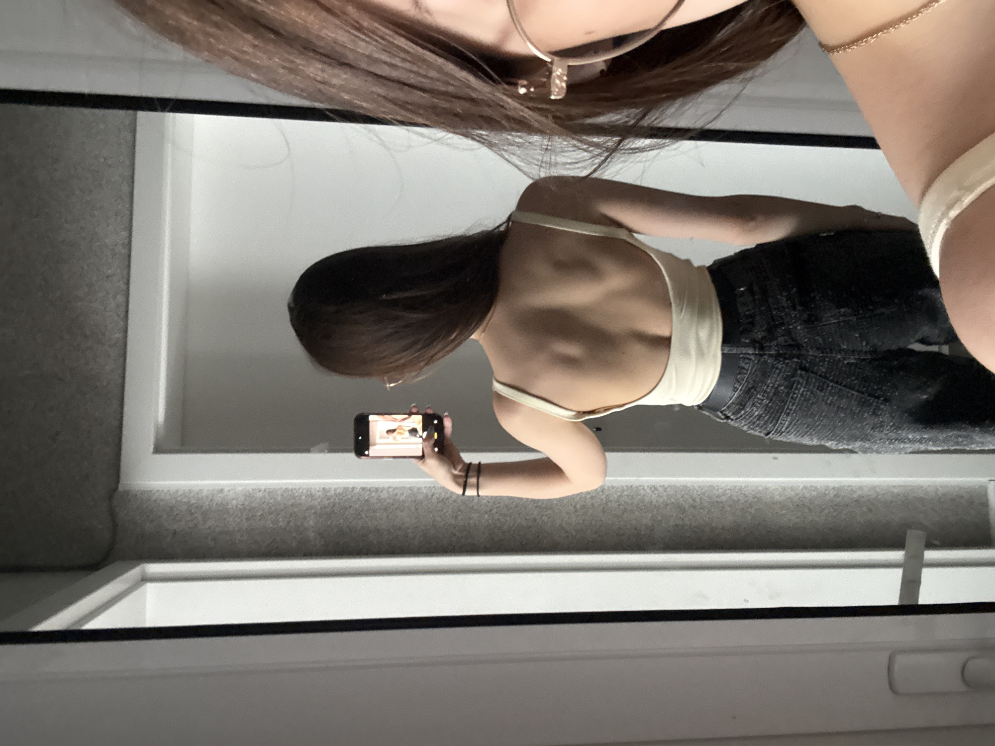 A reflection in a mirror captures a woman holding a smartphone, seemingly taking a photo of herself. She is wearing a light-colored tank top and dark jeans. Part of another woman's head is visible at the top of the mirror frame. The background shows a gray and white interior, possibly a bathroom or bedroom. The location in Sofia, Bulgaria, cannot be determined from the image.