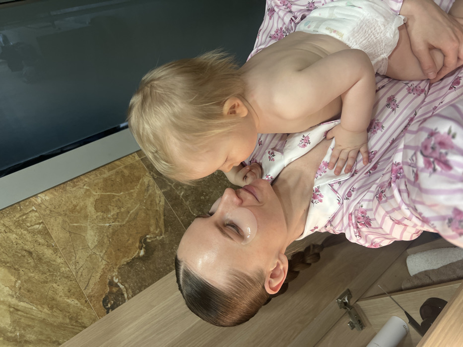 The image shows an adult female holding a young child in an indoor residential setting, likely a bathroom, in the city of Khimki, Russia. The adult female, with her brown hair slicked back and styled into a braid, is wearing a pink and white striped pajama top with a floral pattern. She has translucent hydrogel patches applied beneath both eyes and is looking down at the child with a soft expression. The child, a fair-skinned toddler with blonde hair, is bare-chested and wears a white disposable diaper, which visibly displays the text "Huggies Sun." The child's left hand rests on the woman's chest, and they are engaged in direct eye contact. The background on the left features light-colored wooden cabinetry, with an open compartment revealing a white bottle and folded white towels. The upper and right portions of the background consist of wall tiles with a brown and beige marble pattern, adjacent to a dark, possibly reflective or glass surface on the far right.