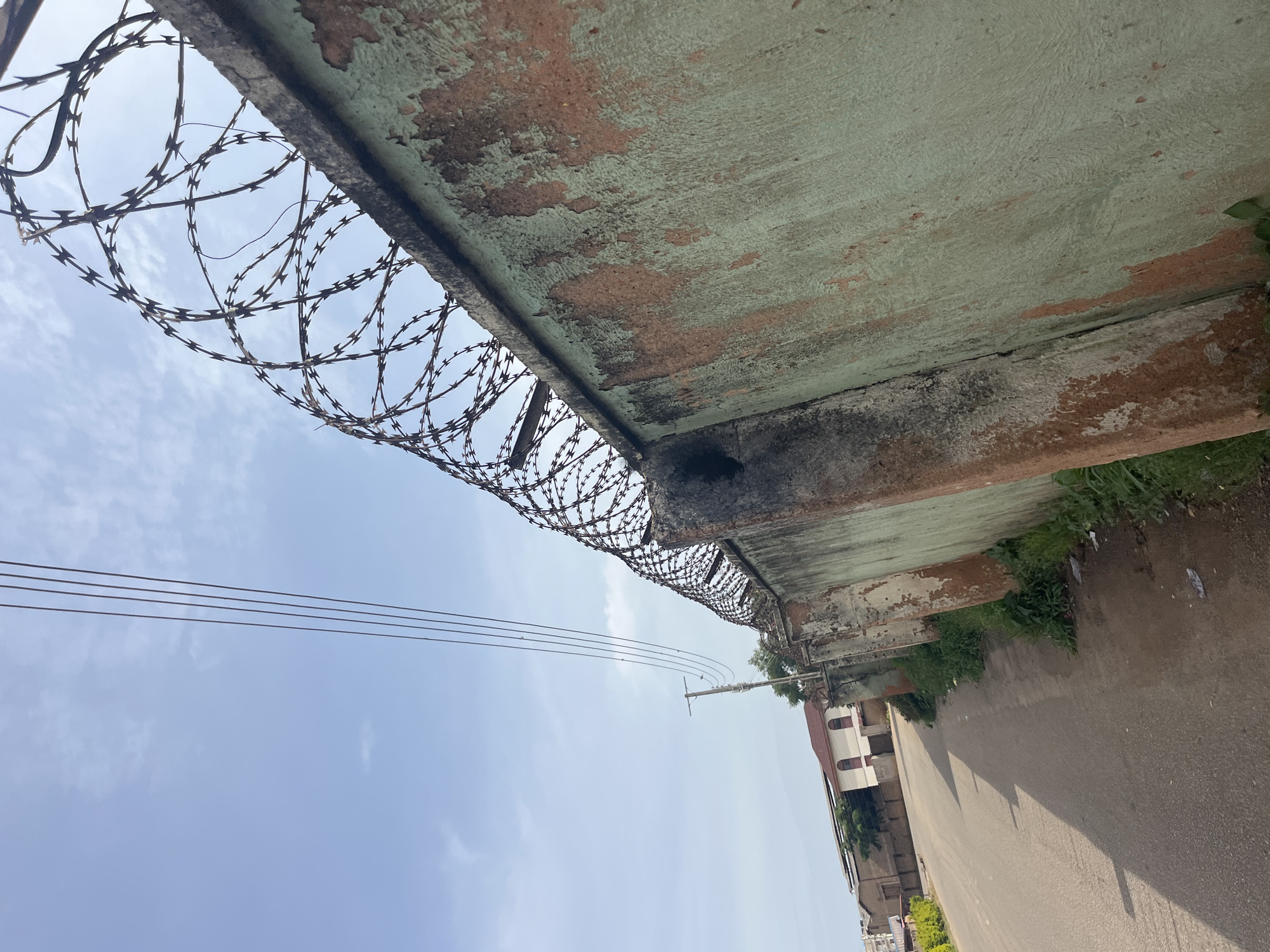 The image captures a street scene in Jos, Nigeria, characterized by a weathered concrete wall topped with barbed wire. The wall, painted a faded green with patches of exposed reddish-brown plaster, runs along the right side of the frame. Above the wall, a tangle of razor wire coils forms a deterrent. In the background, a paved road curves to the left, with a shadow cast across it. Adjacent to the road, on the left side of the frame, are several buildings, including a structure with arched windows and a terracotta-colored roof. A utility pole with electrical wires is visible in the distance. The sky is a light blue with scattered white clouds. The overall scene conveys a sense of an urban environment with some signs of age and wear.