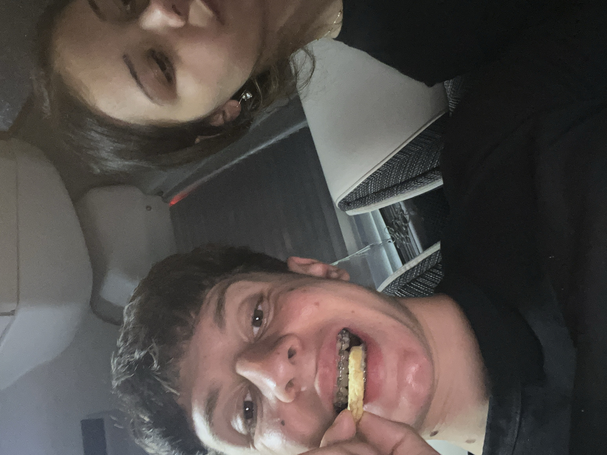 Two individuals are pictured indoors, possibly within a vehicle based on the presence of a headrest and seating in the background. The male subject in the foreground is holding a french fry in his hand, with a portion of it visible between his lips and behind his braces. He is wearing a dark-colored shirt. The female subject is in the background and wearing a dark-colored shirt. The image is angled such that the two individuals are rotated clockwise in the frame.
It is not possible to know if this photo was taken in Popovitsa, Bulgaria.