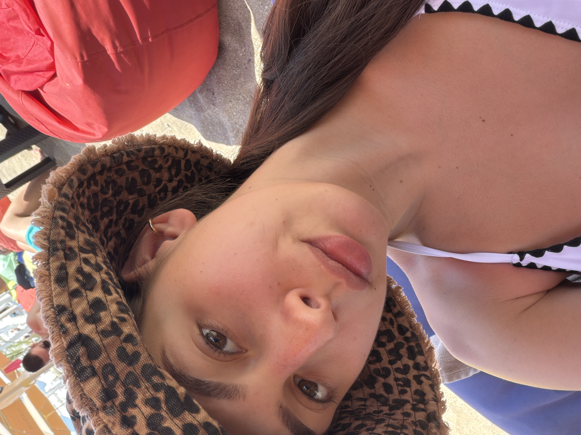 A young woman with dark brown hair is taking a selfie on a beach. She is wearing a leopard-print bucket hat and a white bikini top with black trim. Her skin is tanned and flushed, suggesting she has been in the sun. In the background, a large red beanbag chair is visible, as well as other beachgoers and umbrellas. The scene appears to be a bright, sunny day at the beach, likely in Sozopol, Bulgaria, given the commonality of such scenes along the Bulgarian coast.