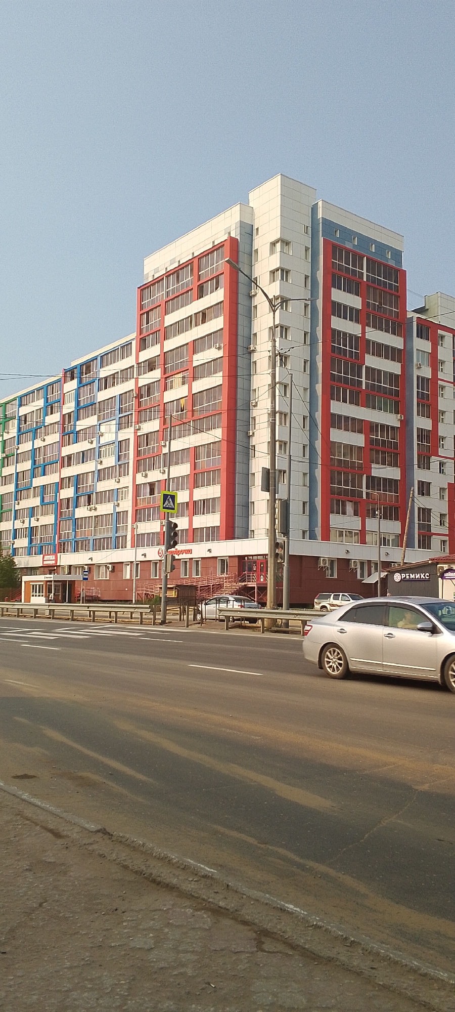 The image depicts a modern, multi-story apartment building with a vibrant facade featuring sections of white, red, and blue. The building has numerous windows and balconies. In the foreground, a street with a pedestrian crossing is visible. Several cars are on the road, including a silver sedan in the immediate foreground and another silver car further back. A bus stop shelter is also present near the building. The scene appears to be on a clear day. Based on the visual cues, specifically the architecture and the presence of Cyrillic script on signage (e.g., "РЕМИКС" and what appears to be "Пятёрочка"), the location is likely within Russia. Without further external information or context, it is not possible to pinpoint the exact location in Yakutsk, Russia.
