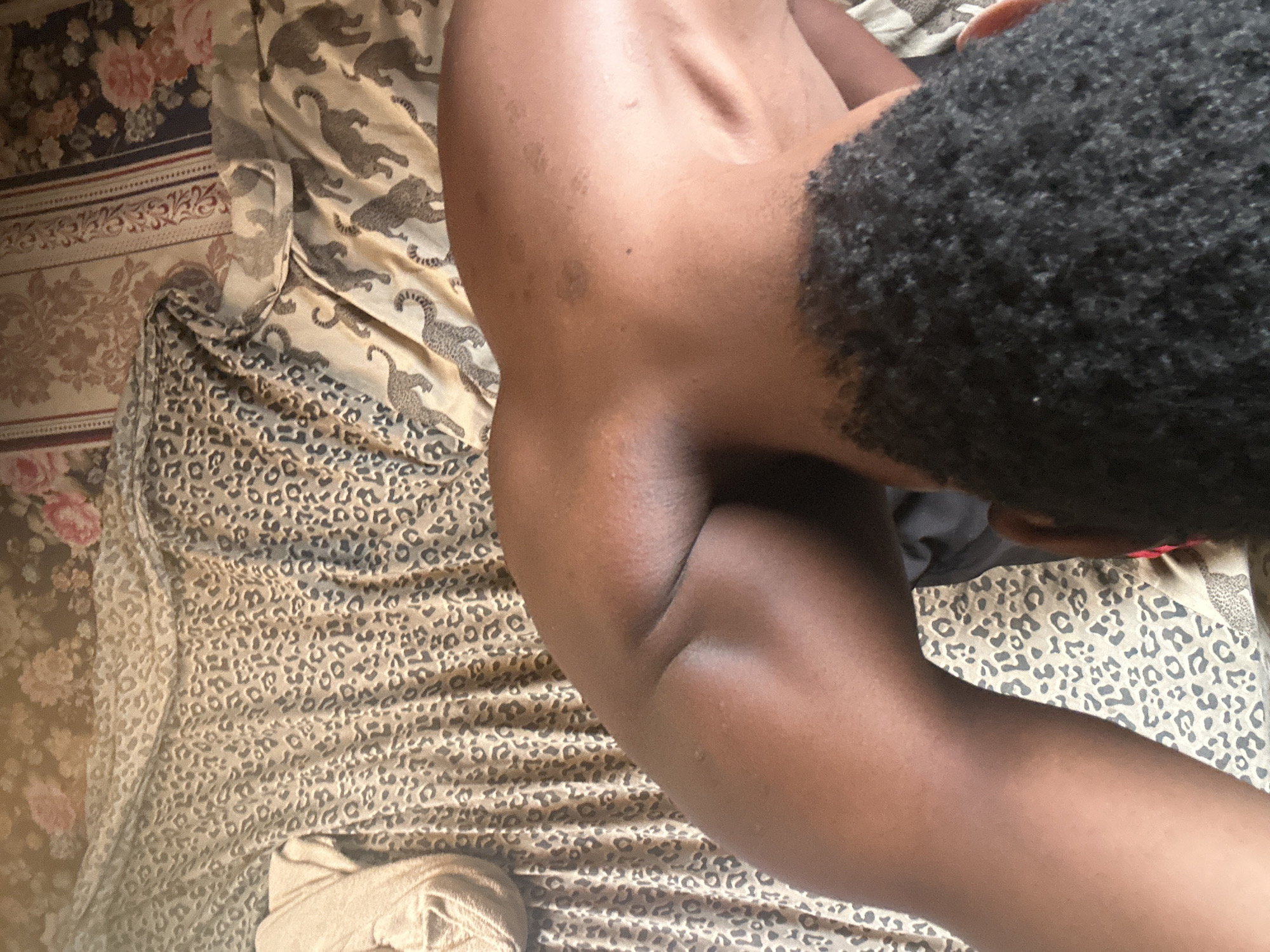 The image captures a close-up, high-angle view of a person lying or resting on a bed. The individual has dark skin and short, tightly curled dark hair, visible on the back of their head and neck. Their bare back, shoulder, and a bent arm are prominently featured, showing some natural skin markings or moles on the upper back.
The setting appears to be a bedroom, characterized by an array of patterned bedding. A dominant light-colored fabric with a dark leopard print covers a large portion of the bed, extending into the foreground. Beneath this, and peeking out from behind the person, is another light-colored fabric featuring stylized, dark cheetah or jaguar figures arranged in a repetitive pattern. Further in the background, a dark blue or black fabric with a vivid floral pattern of red and pink roses is visible, separated by a beige and patterned strip. A light-colored, possibly folded towel or garment is situated on the leopard print bedding in the bottom left corner.
The person's pose suggests relaxation or sleep, with their head turned away from the camera. The lighting indicates it is likely daytime, though no specific light source is visible. There is no discernible text in the image.